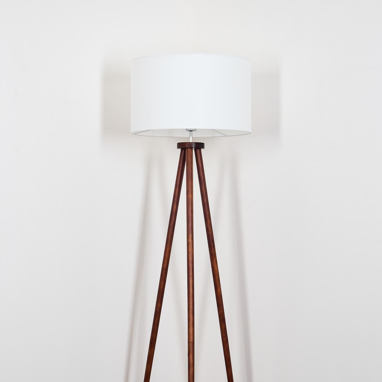 Eine Holz-Stativ-Stehlampe mit weißem Lampenschirm steht vor einer weißen Wand. Die Lampe wirft einen Schatten an die Wand.