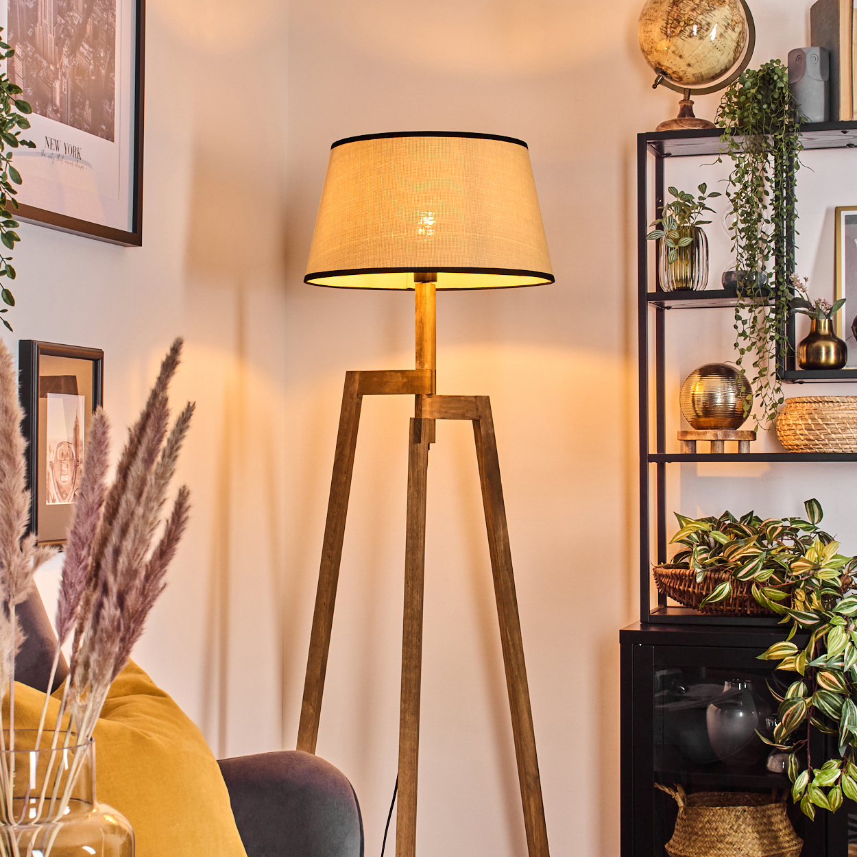 Eine hölzerne Stehlampe mit beigem Lampenschirm. Ein Bücherregal mit Pflanzen und einem Globus. Ein gelbes Kissen und gerahmte Kunst sind sichtbar.