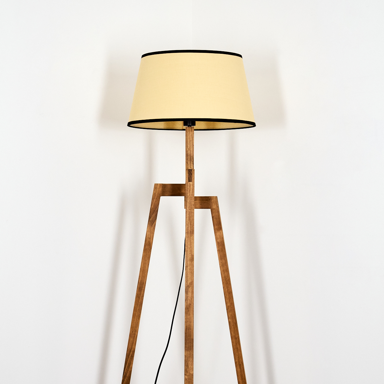 Eine Stehlampe aus Holz mit einem cremefarbenen Lampenschirm mit schwarzer Umrandung steht vor einer weißen Wand. Ein schwarzes Kabel ist sichtbar.