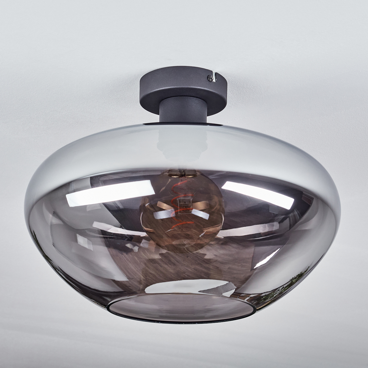 Eine moderne Deckenlampe mit rauchfarbenem Glasschirm und sichtbarer Glühbirne. Die Lampe ist an einer weißen Decke montiert.