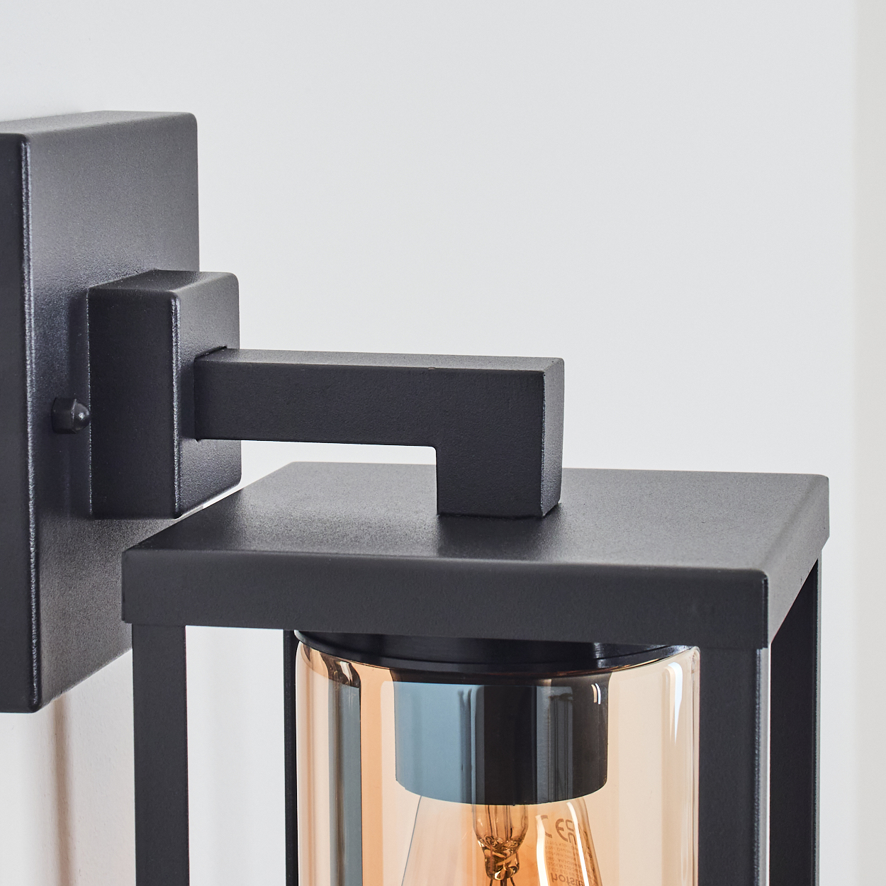 Eine schwarze Außenwandleuchte. Sie hat einen rechteckigen Rahmen mit einem klaren Glaszylinder, der eine bernsteinfarbene Glühbirne enthält. An einer weißen Wand montiert.