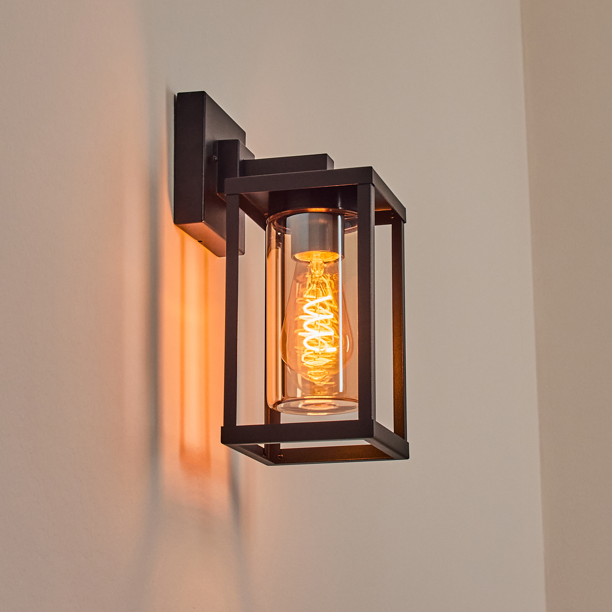 Eine schwarze Wandlampe mit Glaszylinder und einer leuchtenden Glühbirne. An einer hellbeigen Wand montiert. Die Lampe wirft ein warmes Licht.