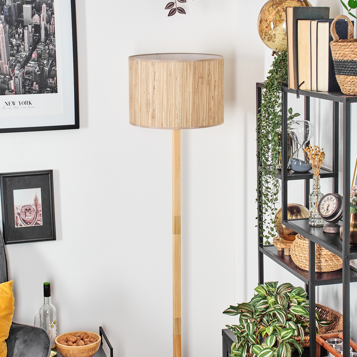 Ein Raum mit einer Stehlampe, einem Bücherregal und gerahmten Bildern. Eine Schale mit Walnüssen und eine Weinflasche stehen auf einem Tisch.
