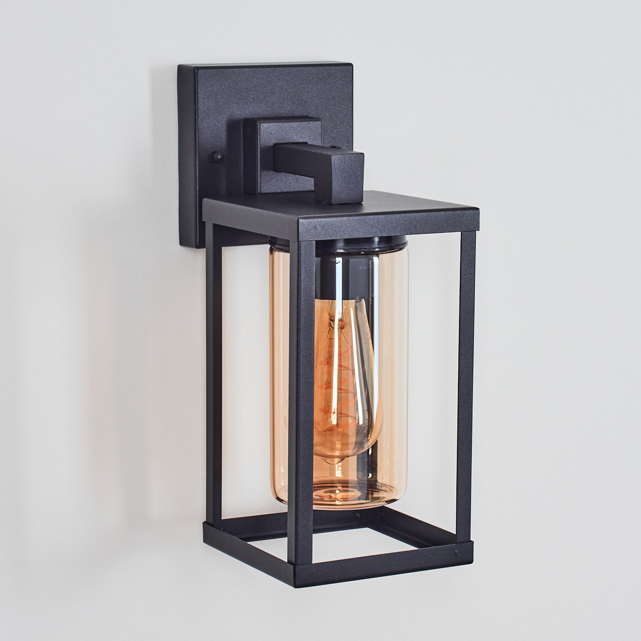 Eine schwarze, an der Wand befestigte Außenleuchte. Sie hat einen rechteckigen Rahmen mit einem zylindrischen, bernsteinfarbenen Glas, das eine Glühbirne umschließt.