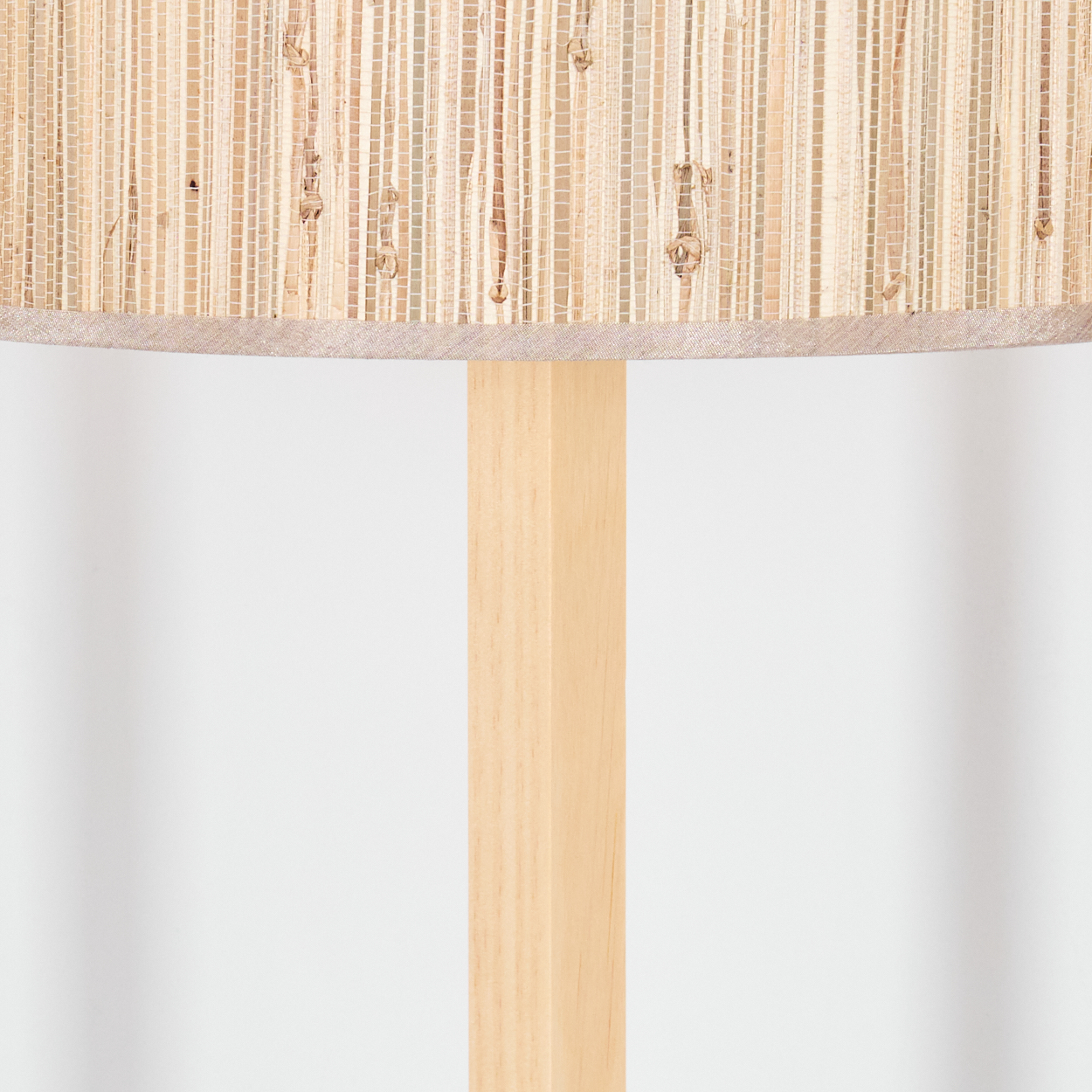Nahaufnahme einer Lampe. Der Lampenschirm ist gewebt, beige. Er sitzt auf einer hellen Holzstütze vor weißem Hintergrund.
