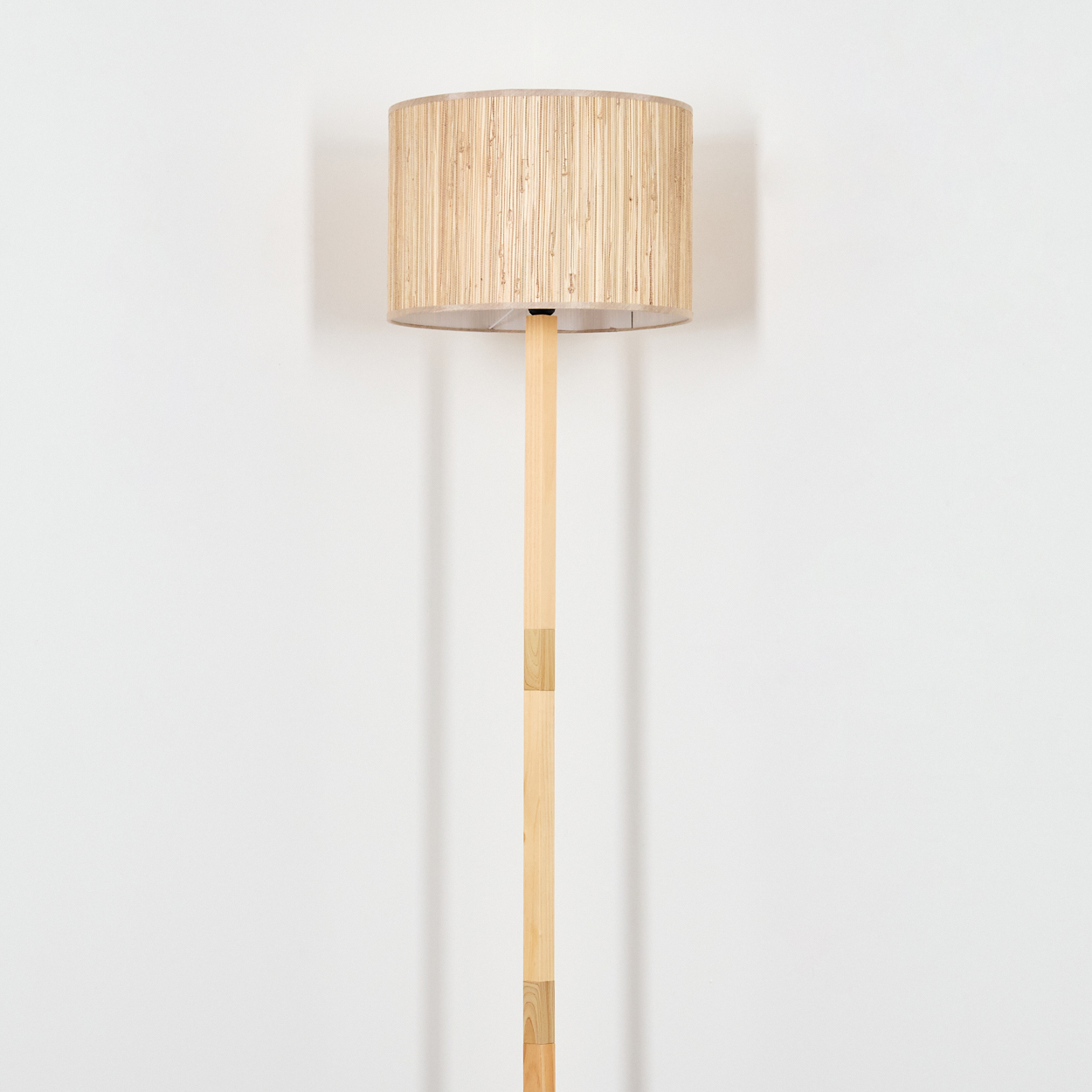 Eine Stehlampe mit einem zylindrischen Lampenschirm aus gewebtem hellbraunem Material. Der Holzpfahl steht vor einer weißen Wand.
