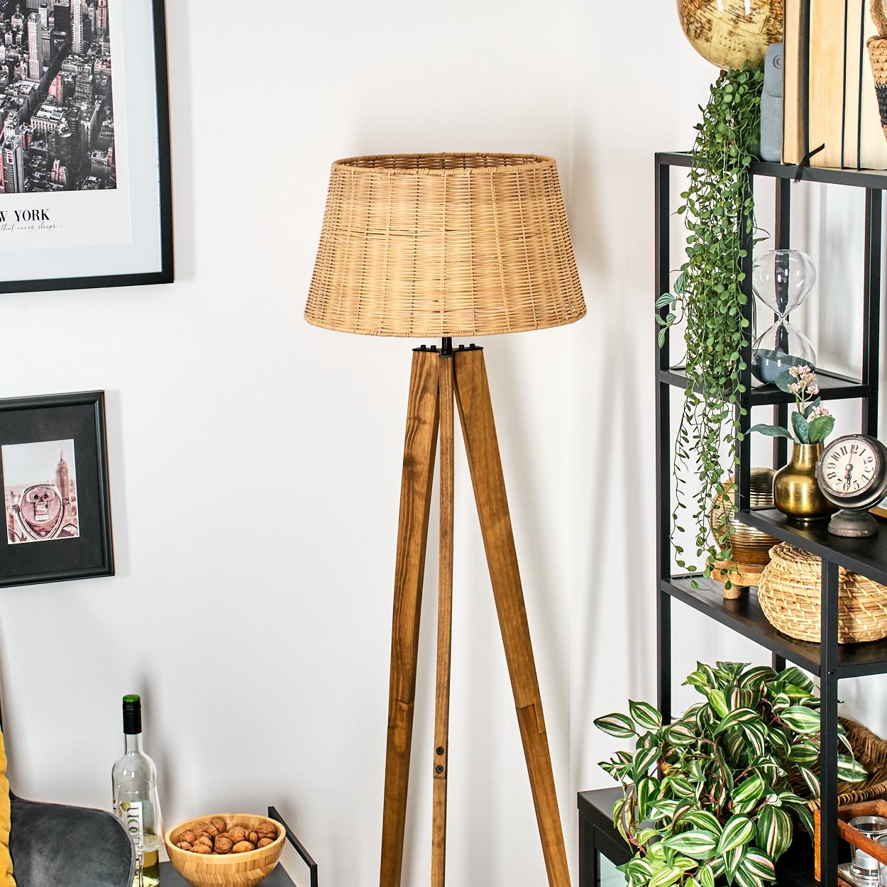 Eine hölzerne Stativ-Stehlampe mit gewebtem Lampenschirm steht neben einem schwarzen Regal mit Pflanzen und Dekoration. An der Wand hängt ein Stadtbild.