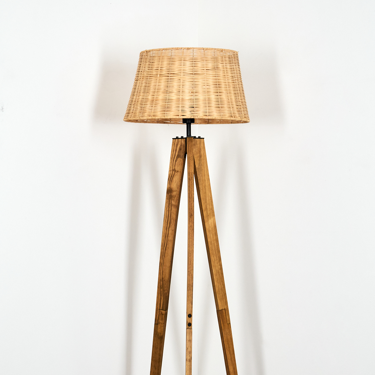 Eine Stehlampe aus Holz mit gewebtem Lampenschirm steht vor einer weißen Wand. Der Sockel ist ein Stativ, der Schirm beige.