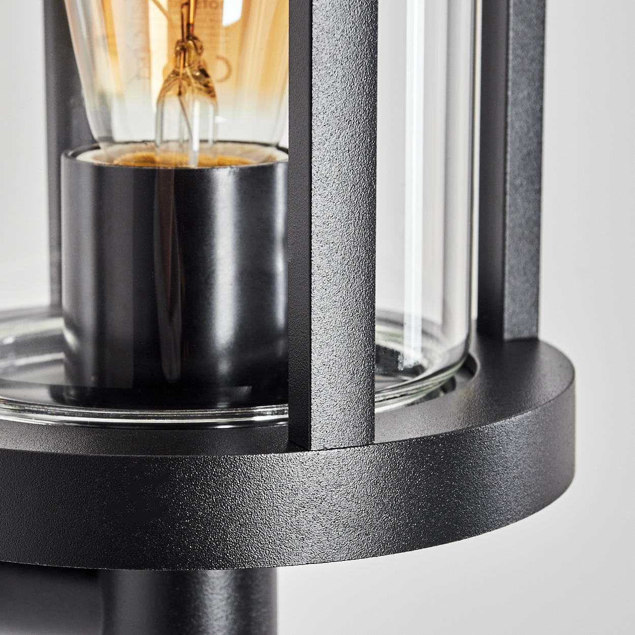 Nahaufnahme einer schwarzen Außenlampe mit Glasgehäuse und sichtbarer Glühbirne. Die Lampe hat eine strukturierte Metalloberfläche.