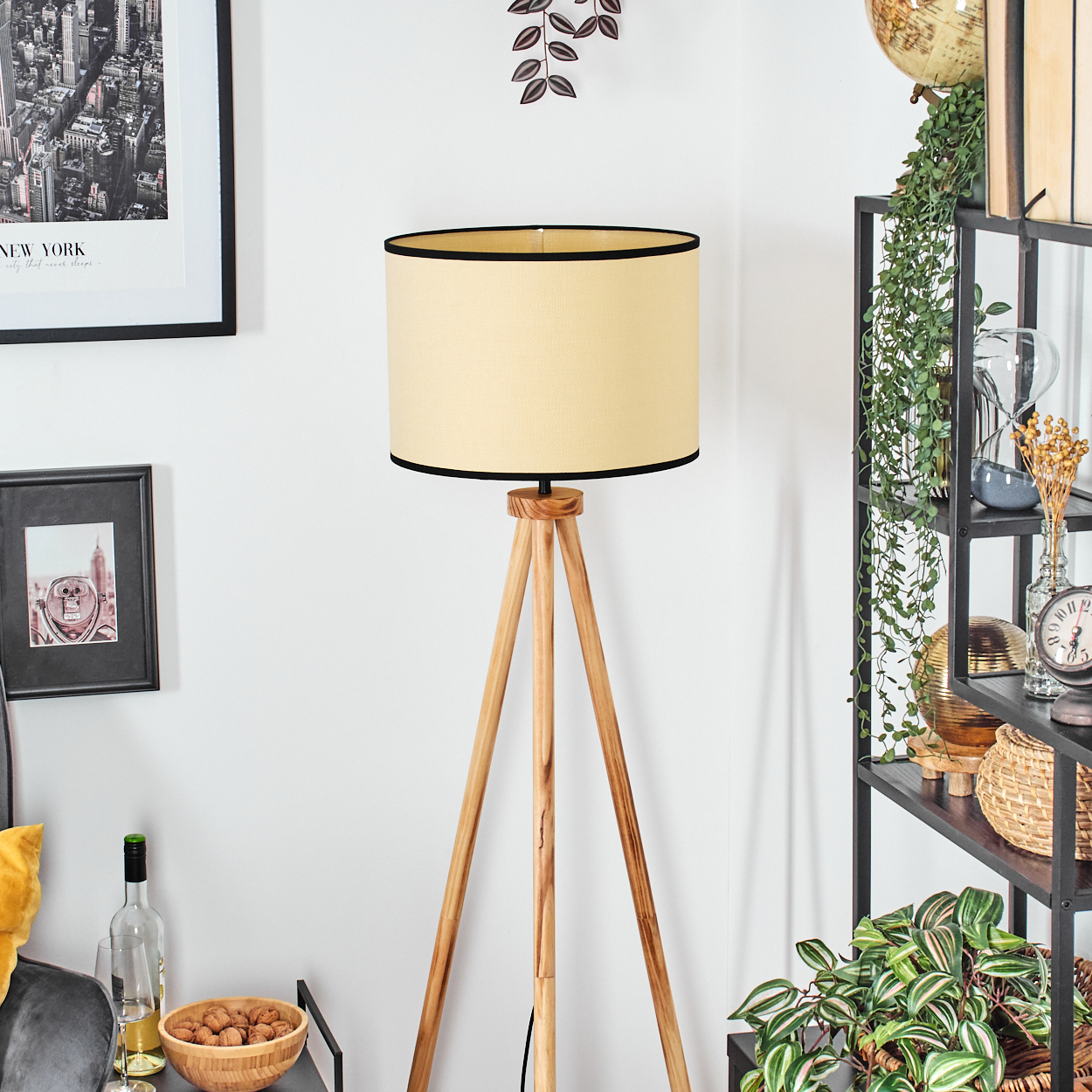 Eine Stehlampe mit Holzstativ und beigem Lampenschirm. Ein Bild mit schwarzem Rahmen und ein Regal mit Deko sind im Hintergrund.