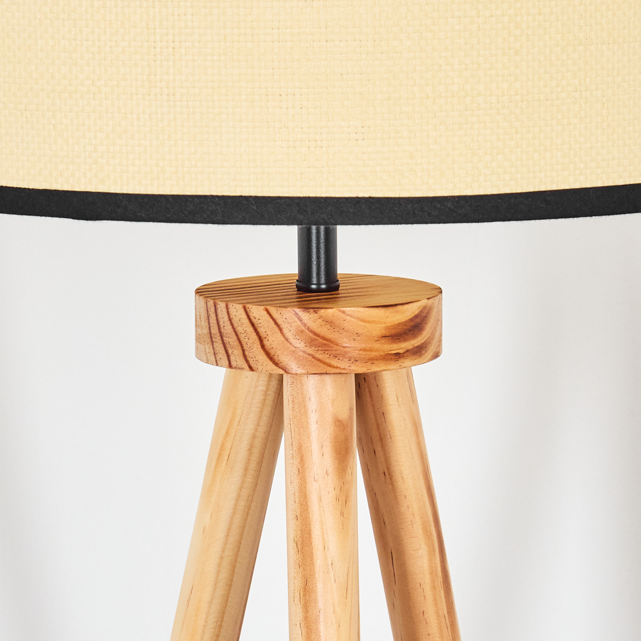 Eine hölzerne Stativlampe. Beigefarbener Lampenschirm mit schwarzem Rand. Helle Holzbeine und -sockel. Schwarze Mittelsäule.