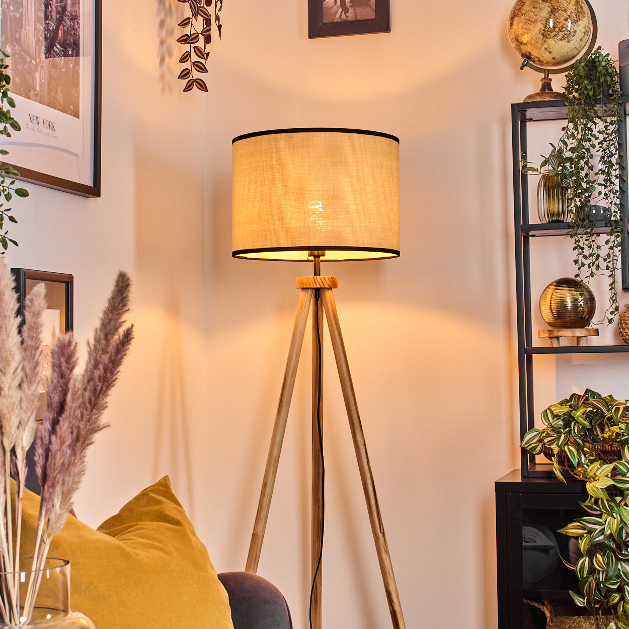 Eine beleuchtete Stehlampe mit beigem Schirm steht in einem Raum. Es gibt ein Sofa mit gelben Kissen. Im Hintergrund ein Regal mit Pflanzen.