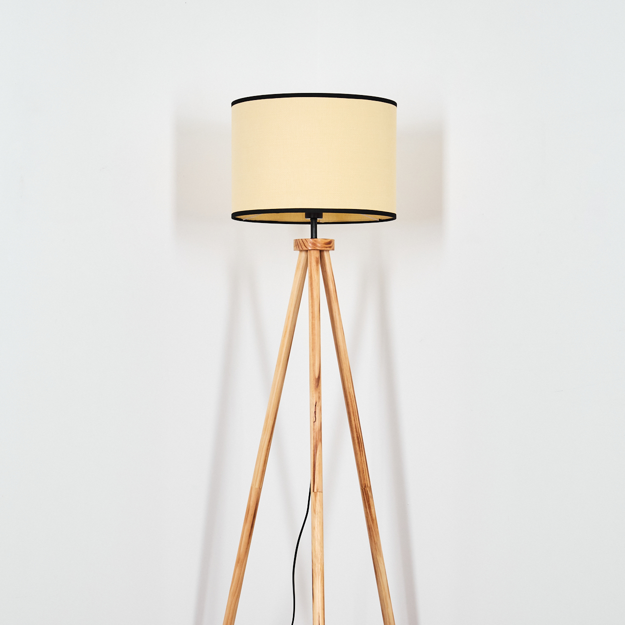 Eine Stehlampe mit Holzstativ, beigem Lampenschirm und schwarzem Rand steht vor einer weißen Wand.