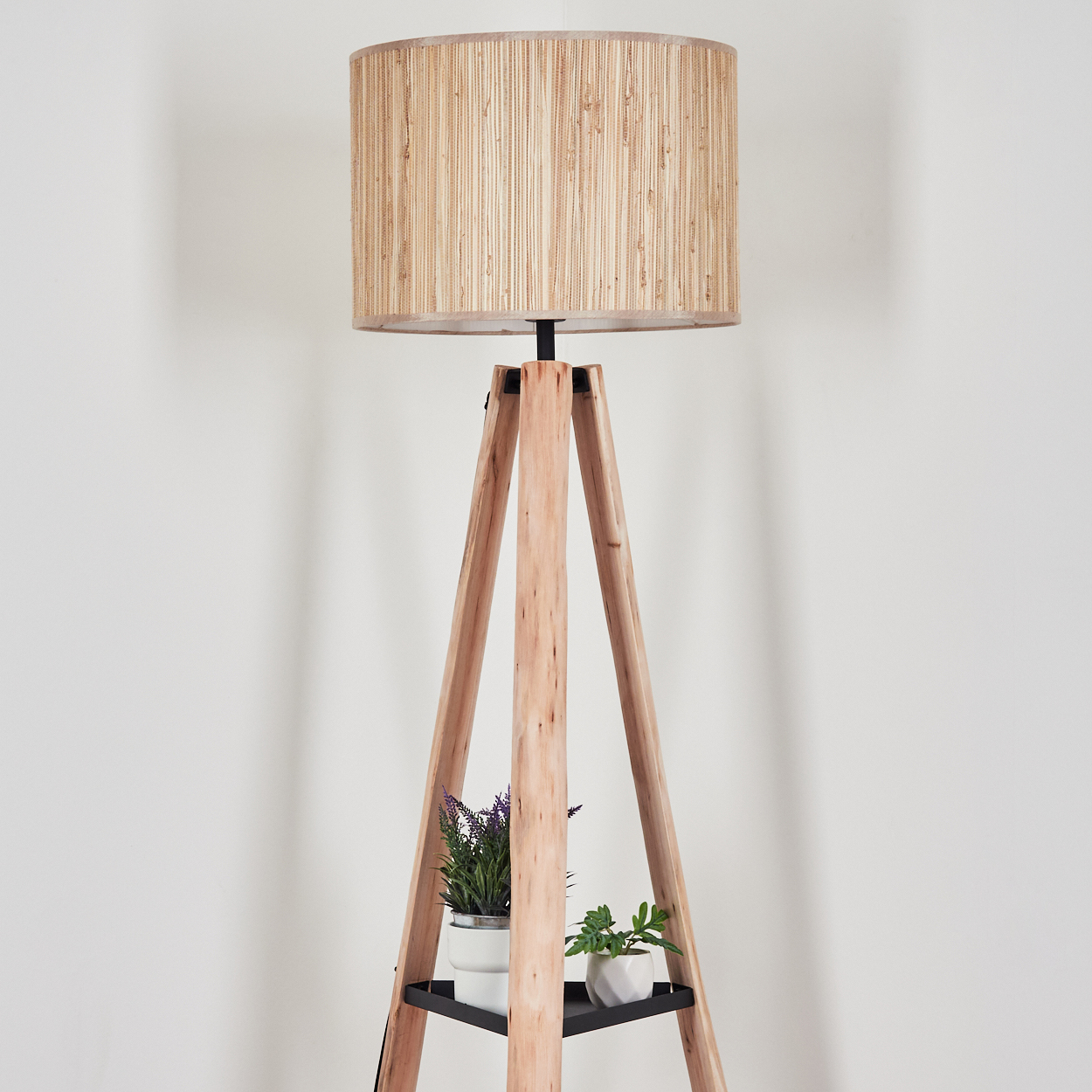 Eine hölzerne Stehlampe mit beigem Lampenschirm. Zwei Topfpflanzen stehen auf einem schwarzen Regal. Der Hintergrund ist eine weiße Wand.