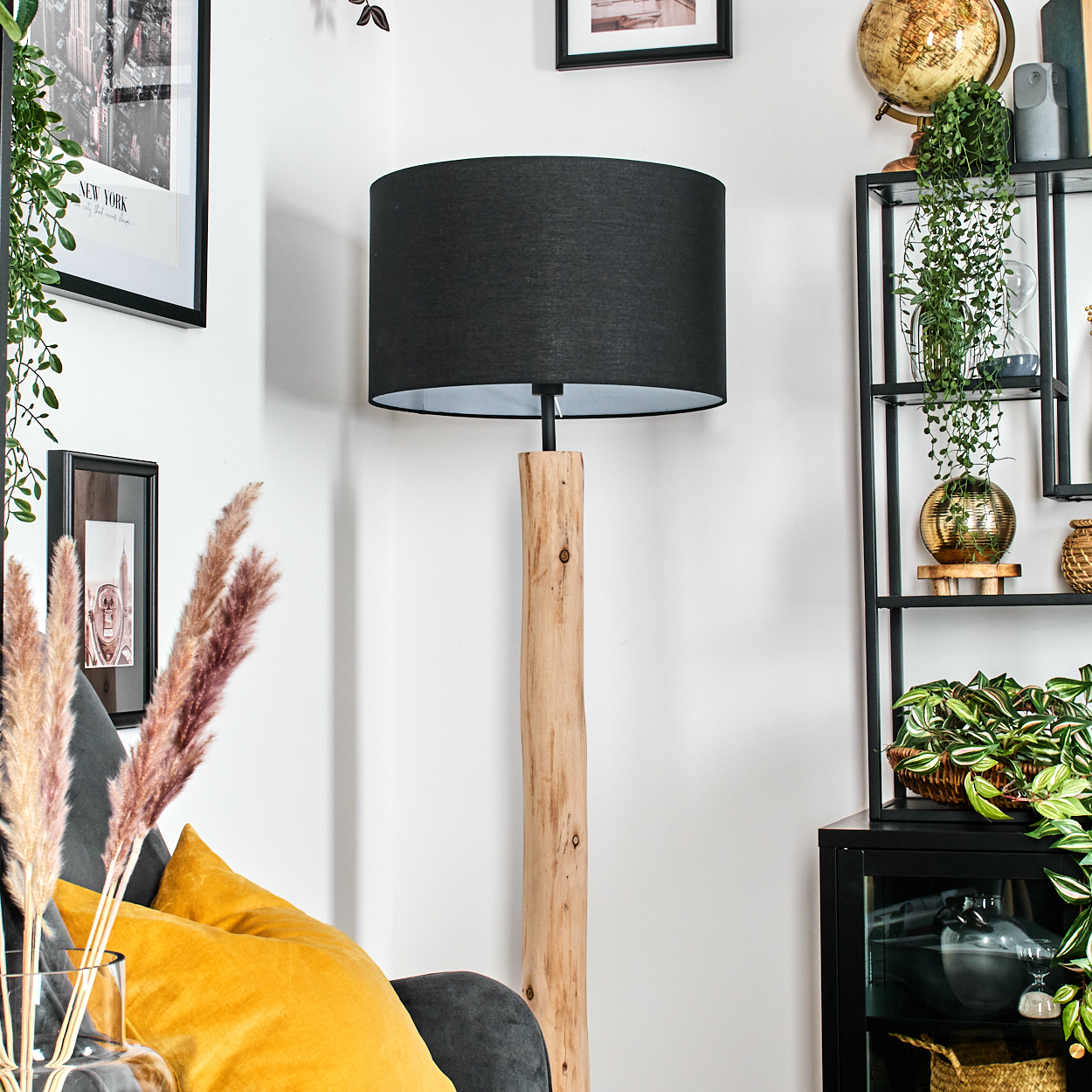 Ein Wohnzimmer mit einer hölzernen Stehlampe, einem schwarzen Lampenschirm und einem Regal mit Pflanzen und Dekoration. Gelbe und graue Kissen auf dem Sofa.