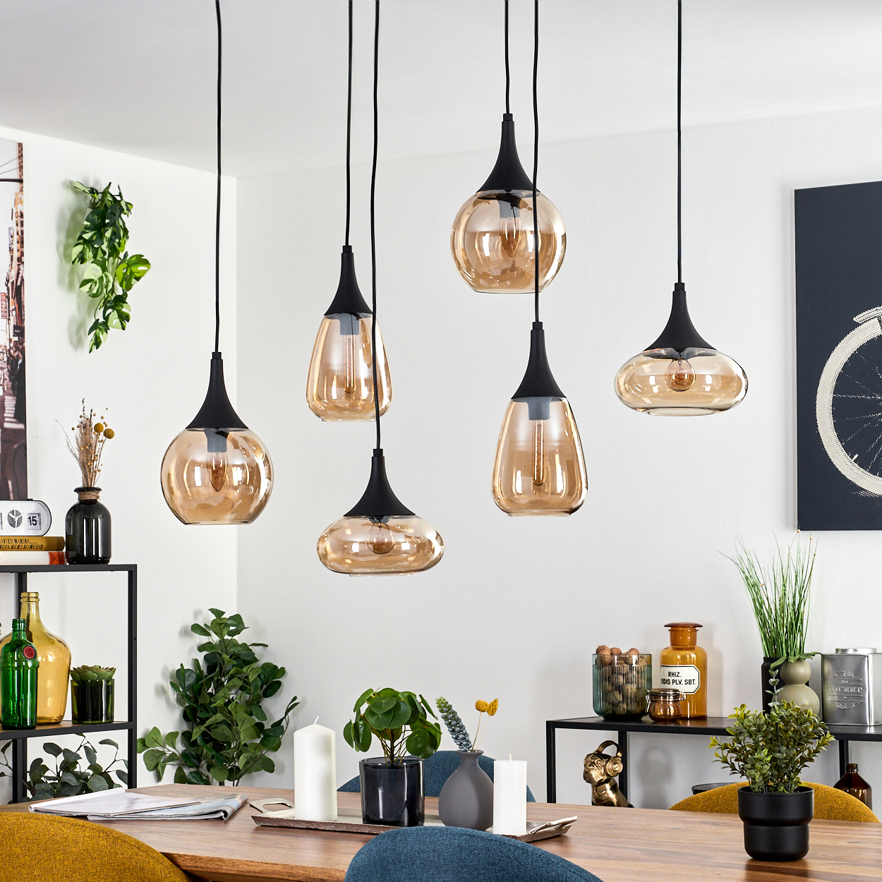 Sechs bernsteinfarbene Glas-Pendelleuchten hängen über einem Holztisch. Pflanzen, Kerzen und Dekoration stehen auf dem Tisch und in Regalen.