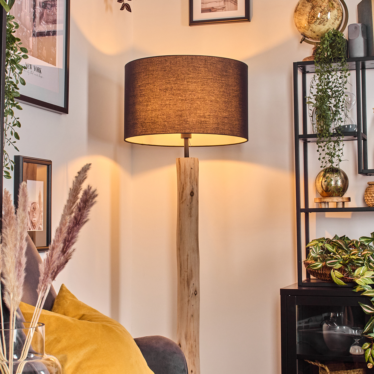 Eine Stehlampe aus Holz mit schwarzem Lampenschirm. Ein gelbes Kissen und ein Regal mit Pflanzen sind ebenfalls zu sehen.