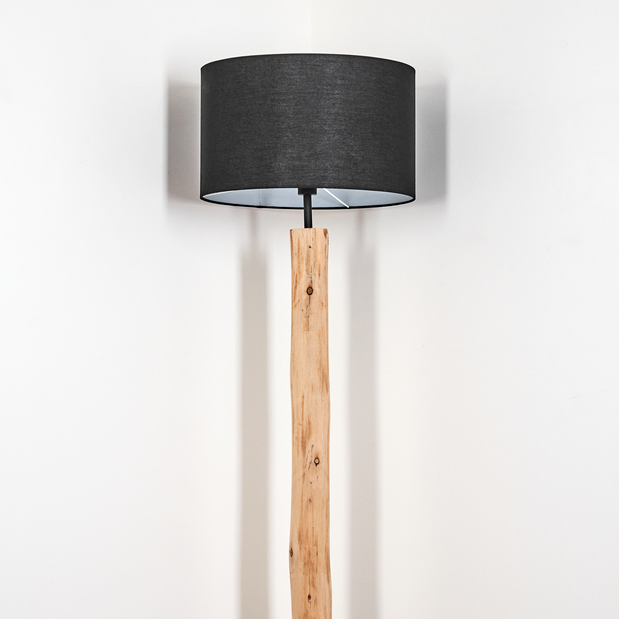 Eine Stehlampe mit schwarzem Lampenschirm und Holzfuß steht vor einer weißen Wand. Der Schirm wirft einen Schatten.
