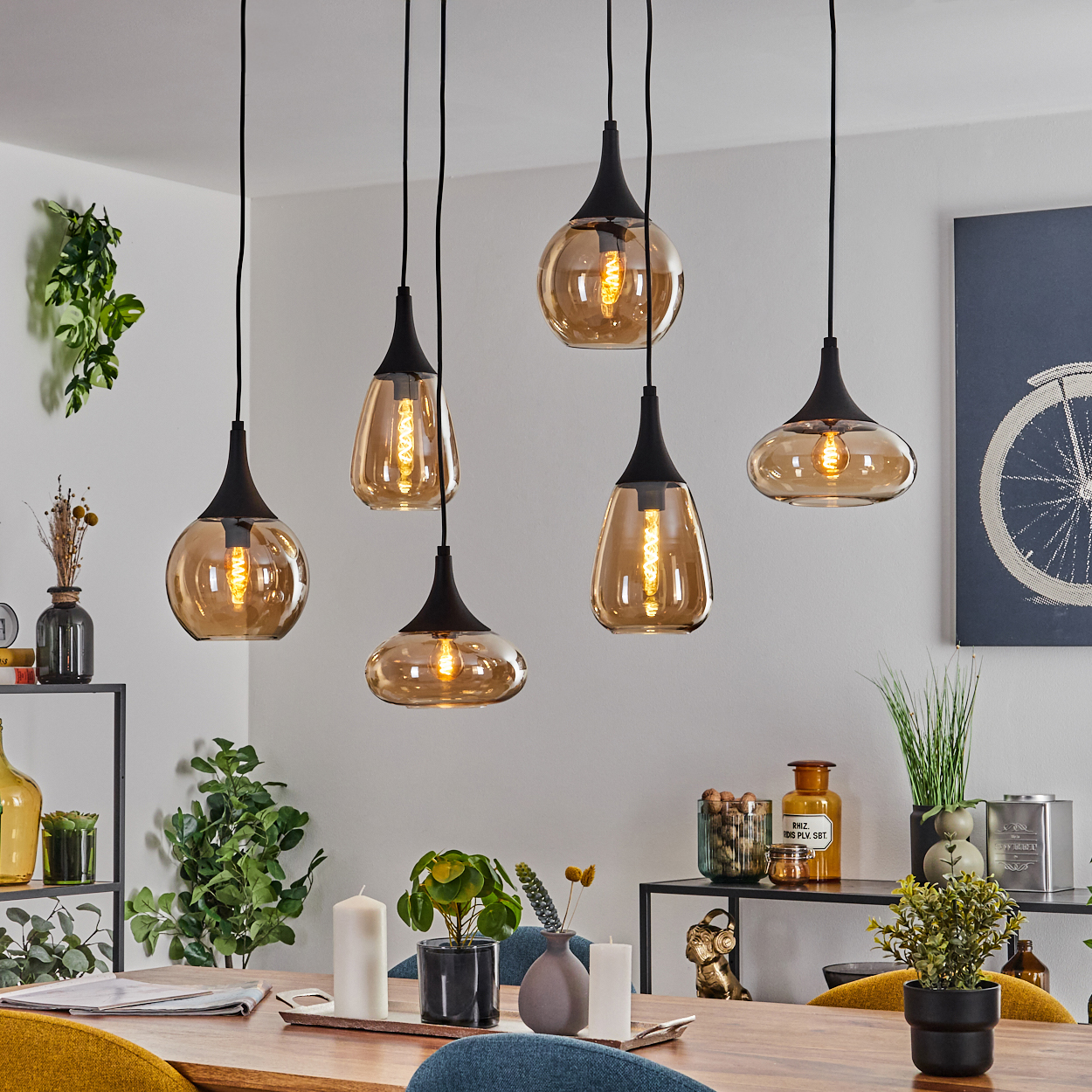 Sechs hängende bernsteinfarbene Glas-Pendelleuchten über einem Holzesstisch mit Pflanzen und Dekoration. Ein Gemälde mit einem Fahrradrad an der Wand.