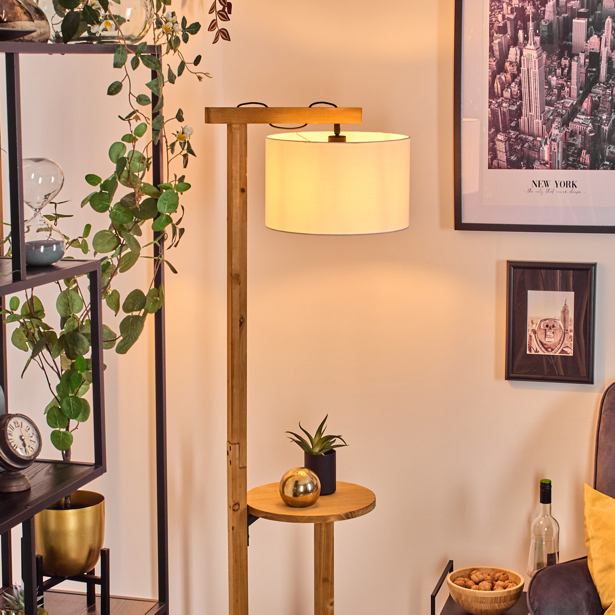 Eine hölzerne Stehlampe mit Lampenschirm. Ein Regal mit Pflanzen und Dekoration. Ein New York-Bild an der Wand. Weitere Deko auf dem Beistelltisch.