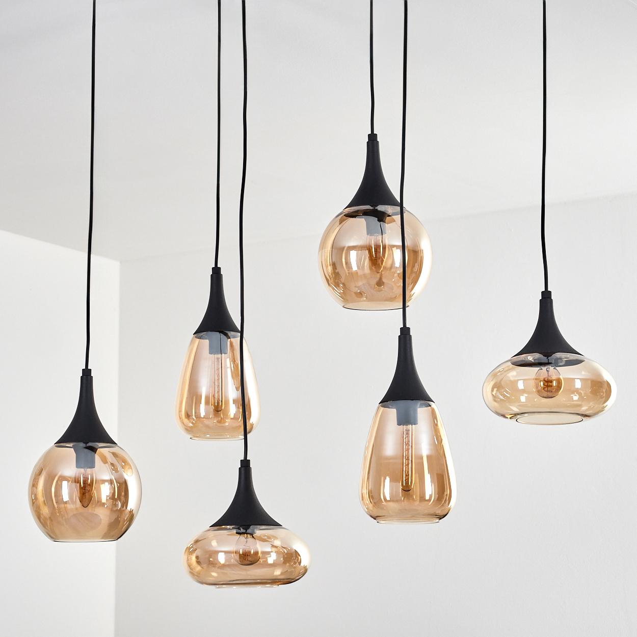 Mehrere hängende Pendelleuchten mit bernsteinfarbenen Glasschirmen und schwarzen Armaturen vor einer weißen Wand.