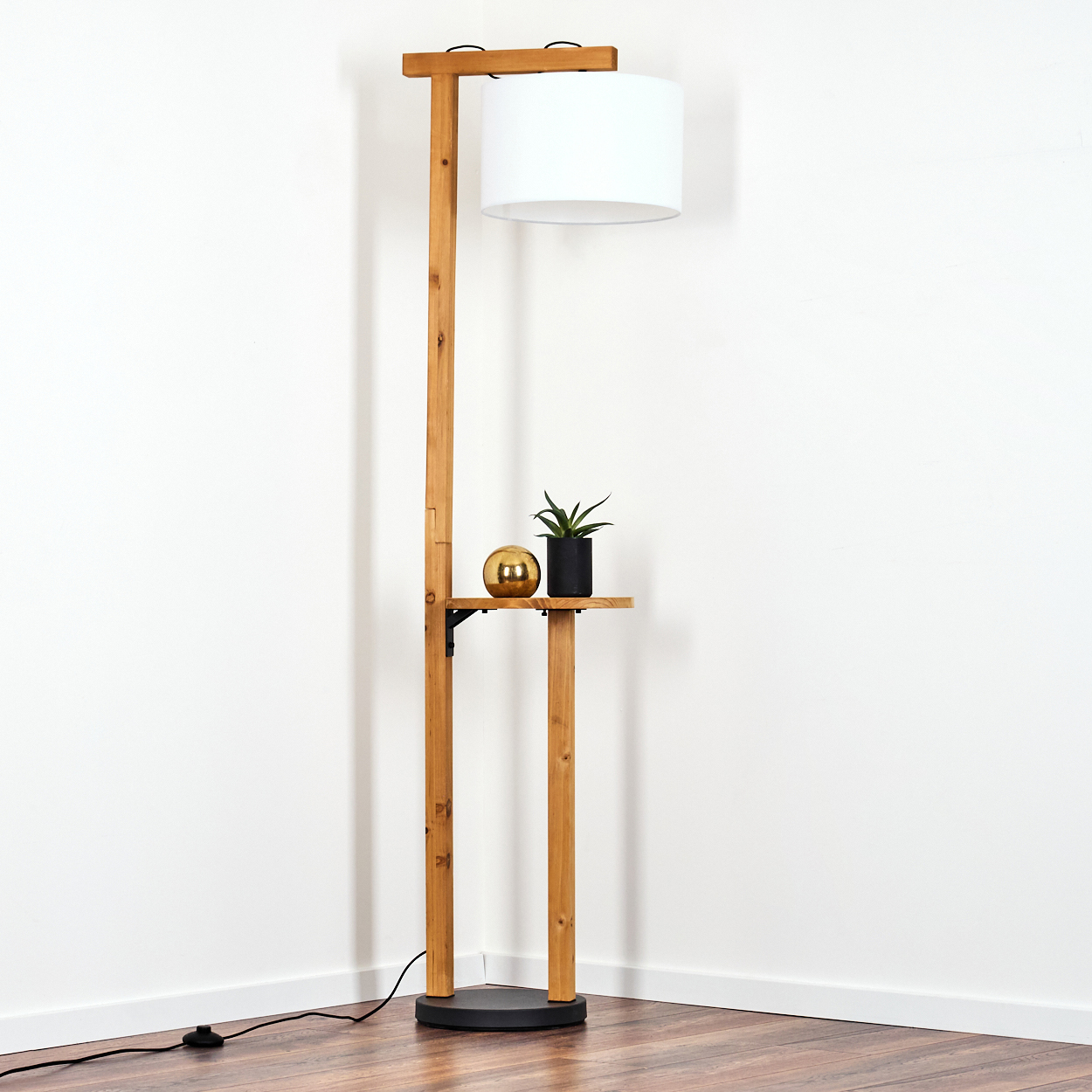 Eine Stehlampe aus Holz mit weißem Lampenschirm, ein kleines Regal mit goldener Kugel und Pflanze, vor einer weißen Wand.