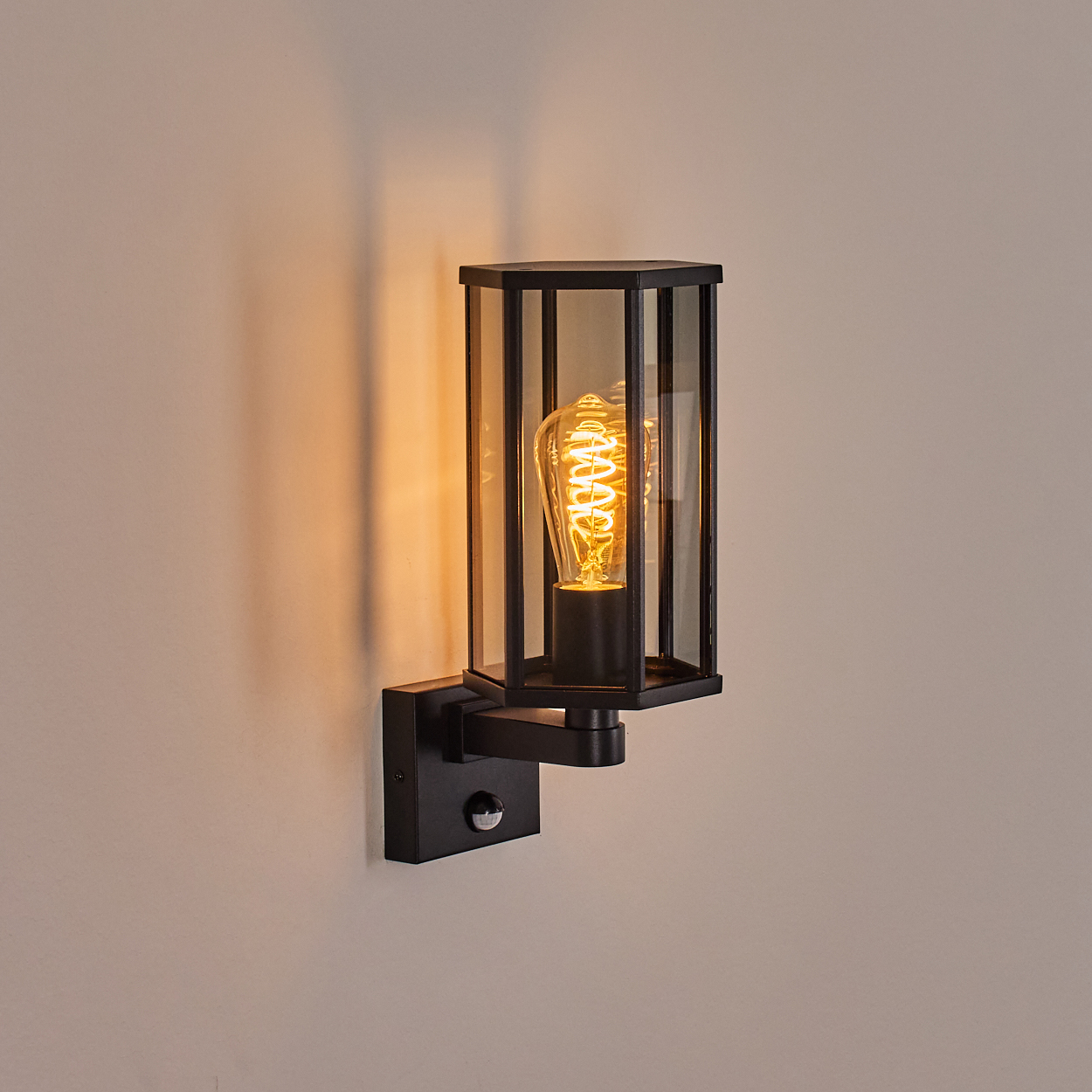 Eine schwarze Wandleuchte mit Glasgehäuse und leuchtender Glühbirne. Die Wand ist hellbeige.