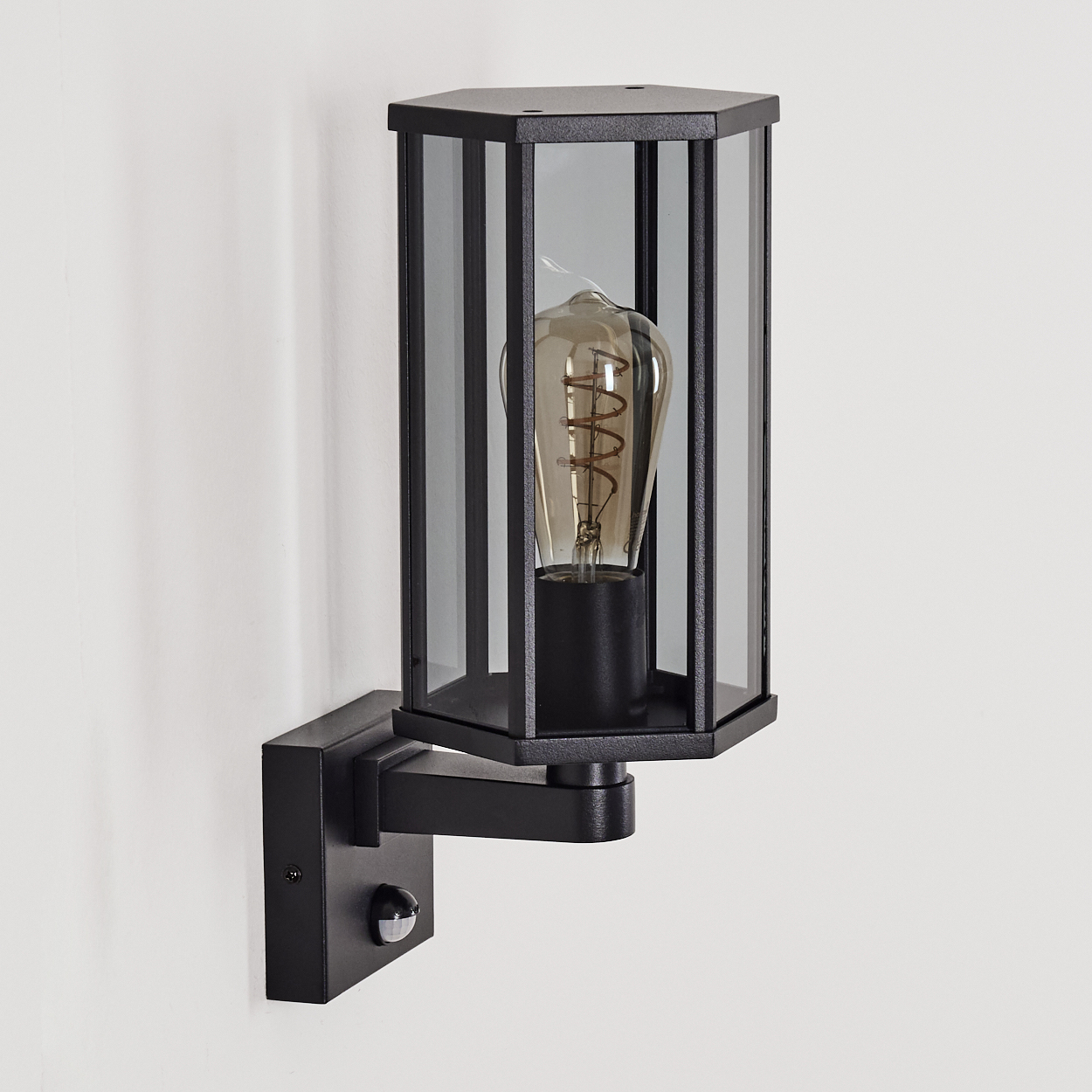 Schwarze Außenwandleuchte mit sechseckigem Schirm, Glasscheiben und einer sichtbaren Glühbirne, an einer weißen Wand montiert.