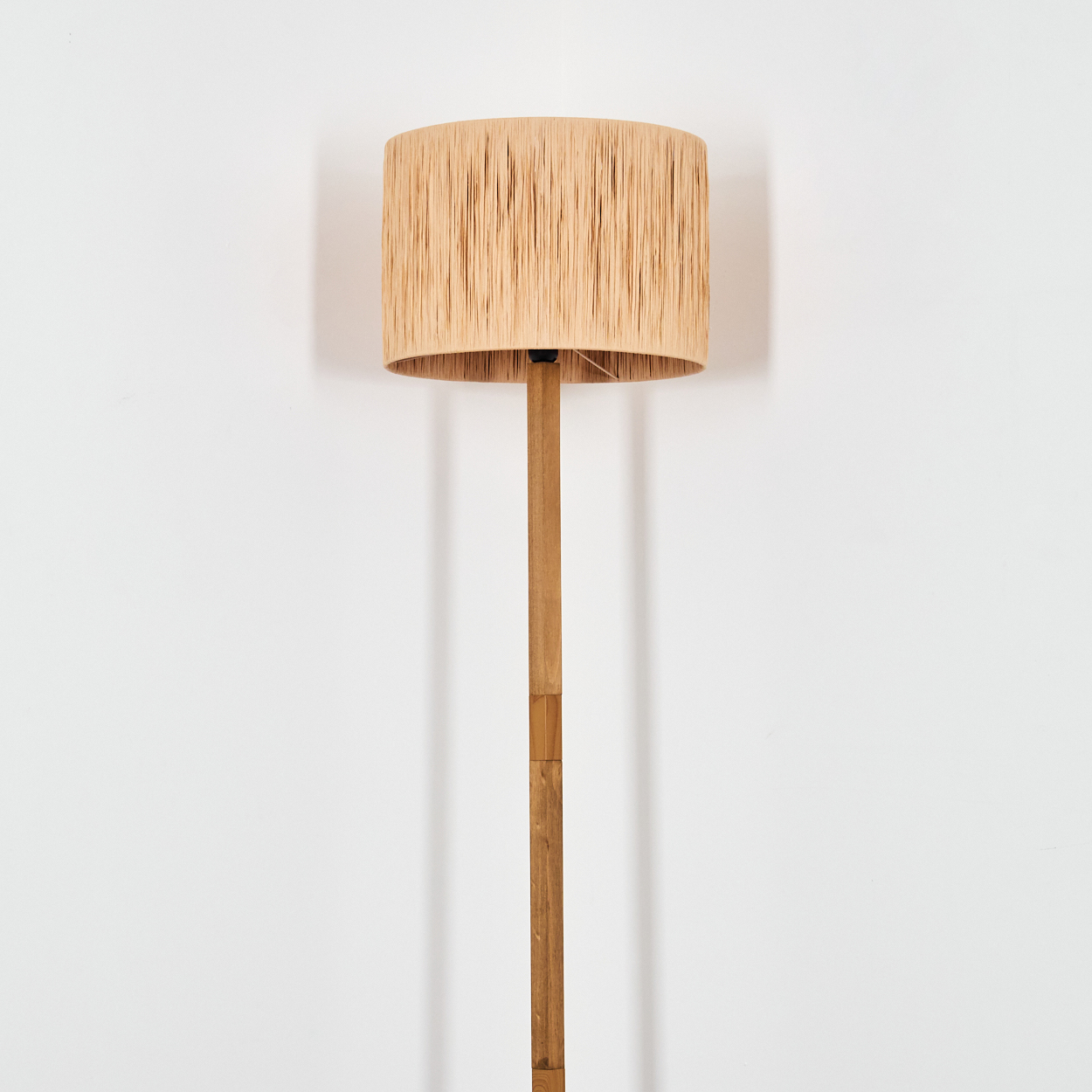 Eine Stehlampe mit beigem, gewebtem Lampenschirm und Holzpfahl. Der Hintergrund ist eine schlichte, weiße Wand.