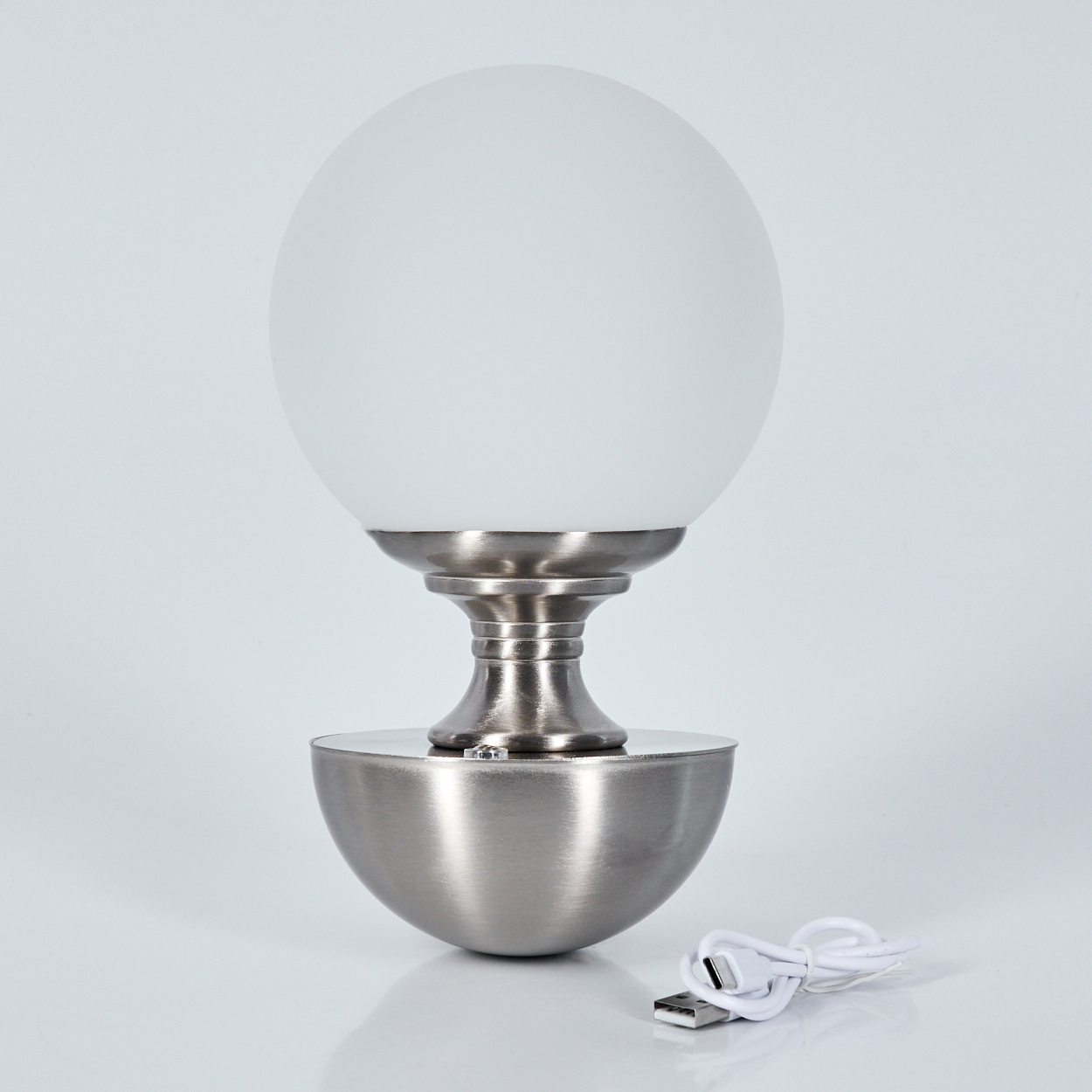 Eine moderne Lampe mit weißem Kugelschirm, silbernem Sockel und USB-Kabel. Die Lampe steht auf einer weißen Oberfläche.
