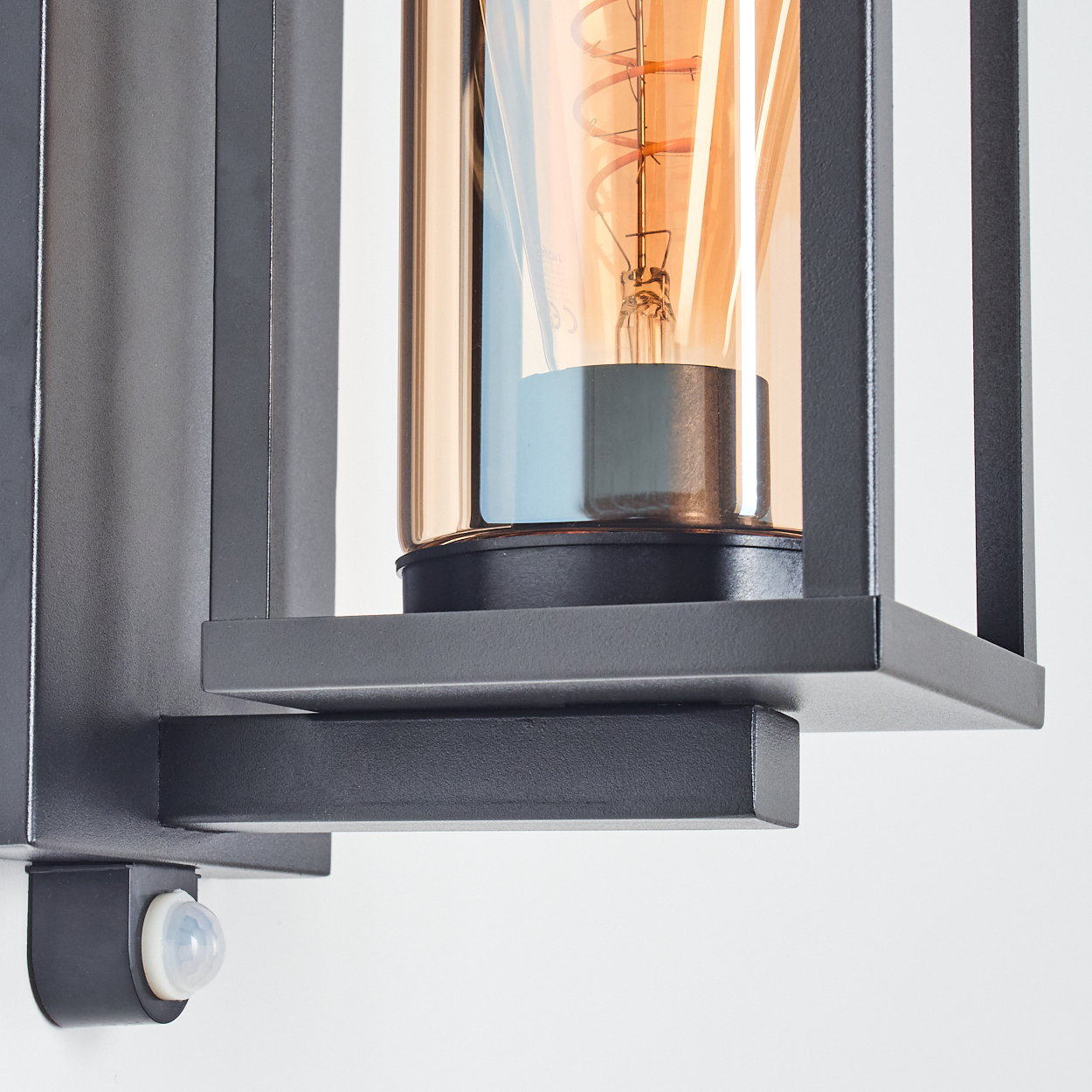 Eine schwarze Außenleuchte mit einer warm getönten Glühbirne, die in einem Metallrahmen eingeschlossen ist, und einem Bewegungssensor.