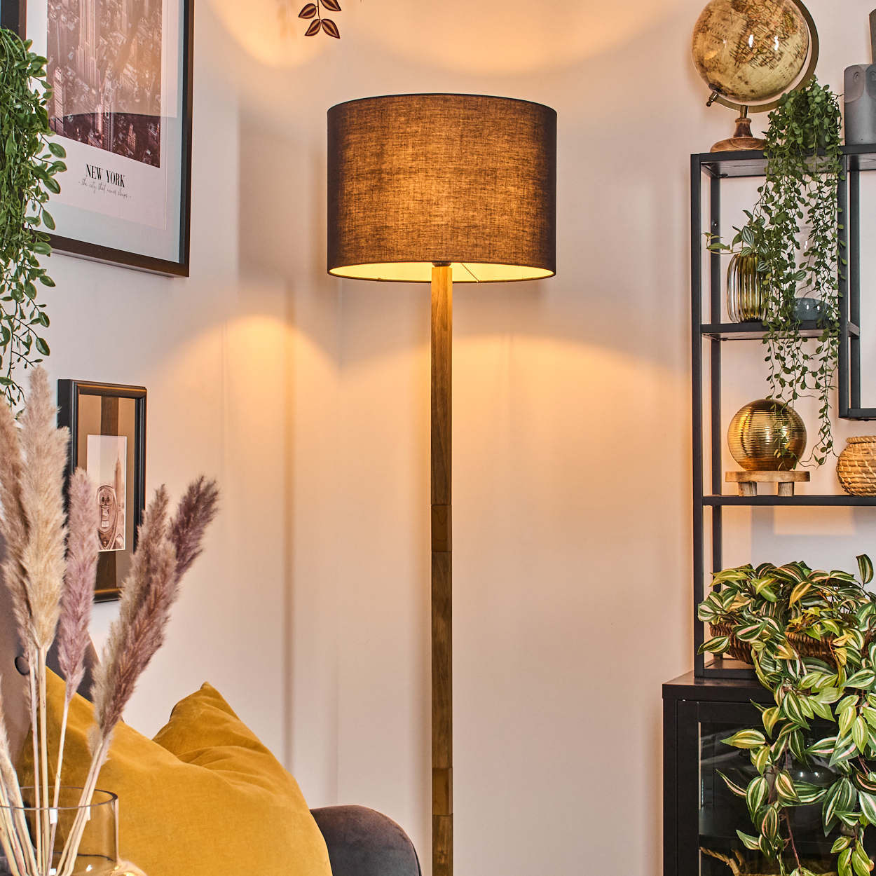 Eine beleuchtete Stehlampe mit dunklem Schirm steht neben einem Regal mit Globus und Pflanzen. Ein Bild von New York hängt an der Wand.