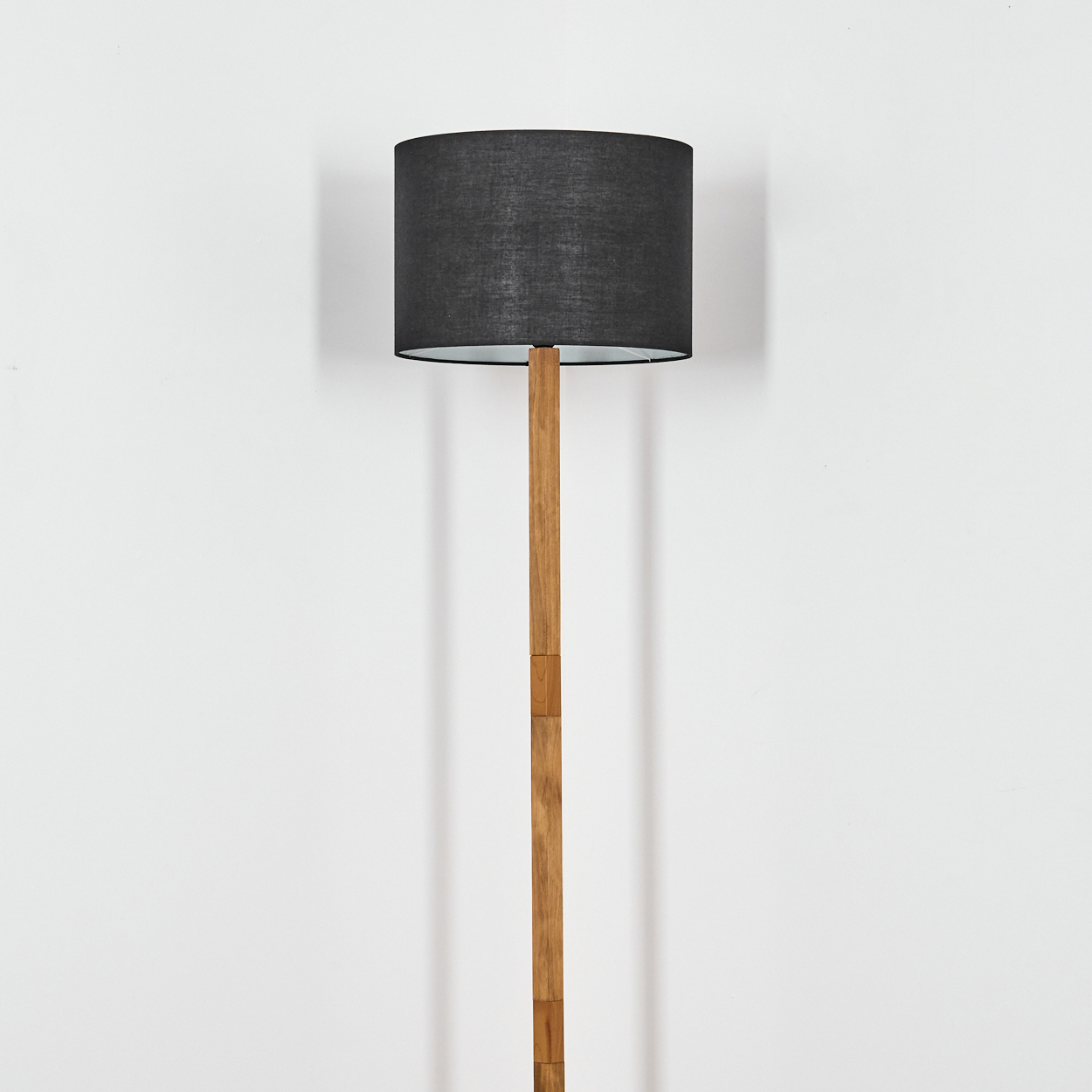Eine Stehlampe mit dunklem Lampenschirm und Holzständer. Sie steht vor einer weißen Wand. Die Lampe ist zentriert.
