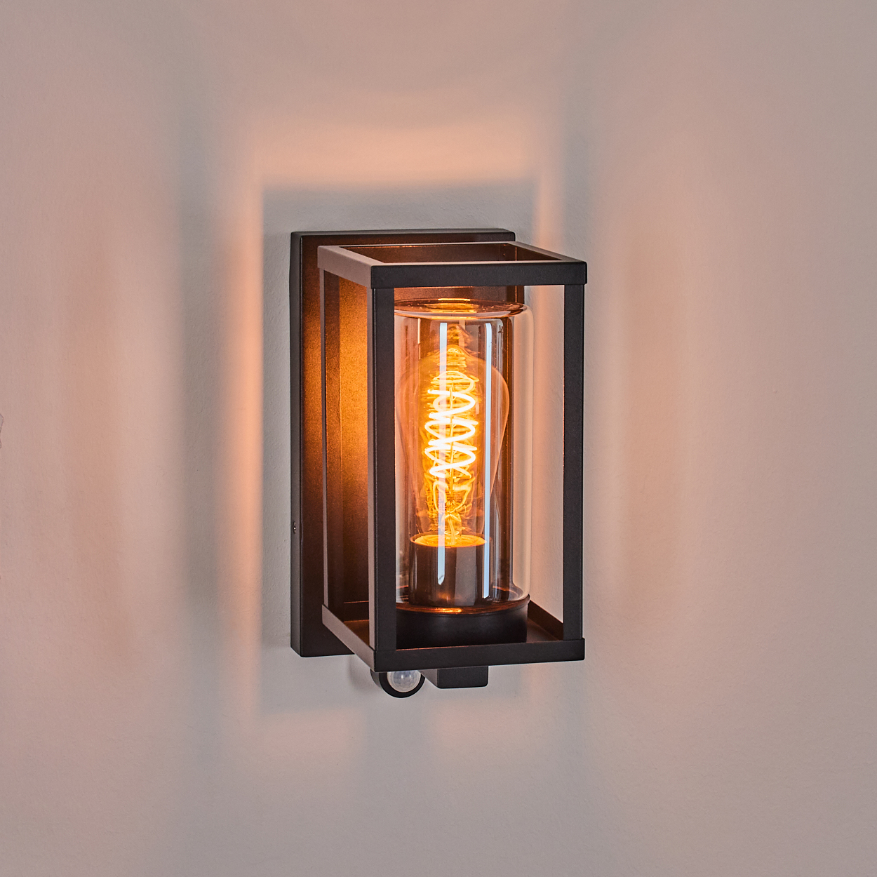 Eine schwarze Außenwandleuchte mit einer warmgelben Glühbirne in einem Glaszylinder, an einer weißen Wand befestigt.