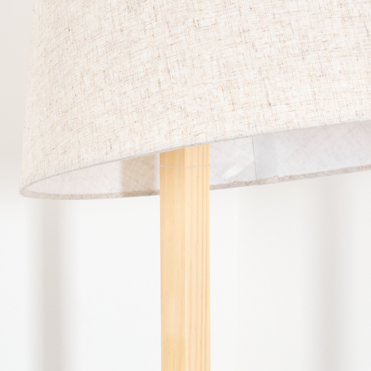 Nahaufnahme einer Stehlampe. Sie hat einen beigefarbenen Lampenschirm und einen hellen Holzrahmen. Der Hintergrund ist eine schlichte weiße Wand.
