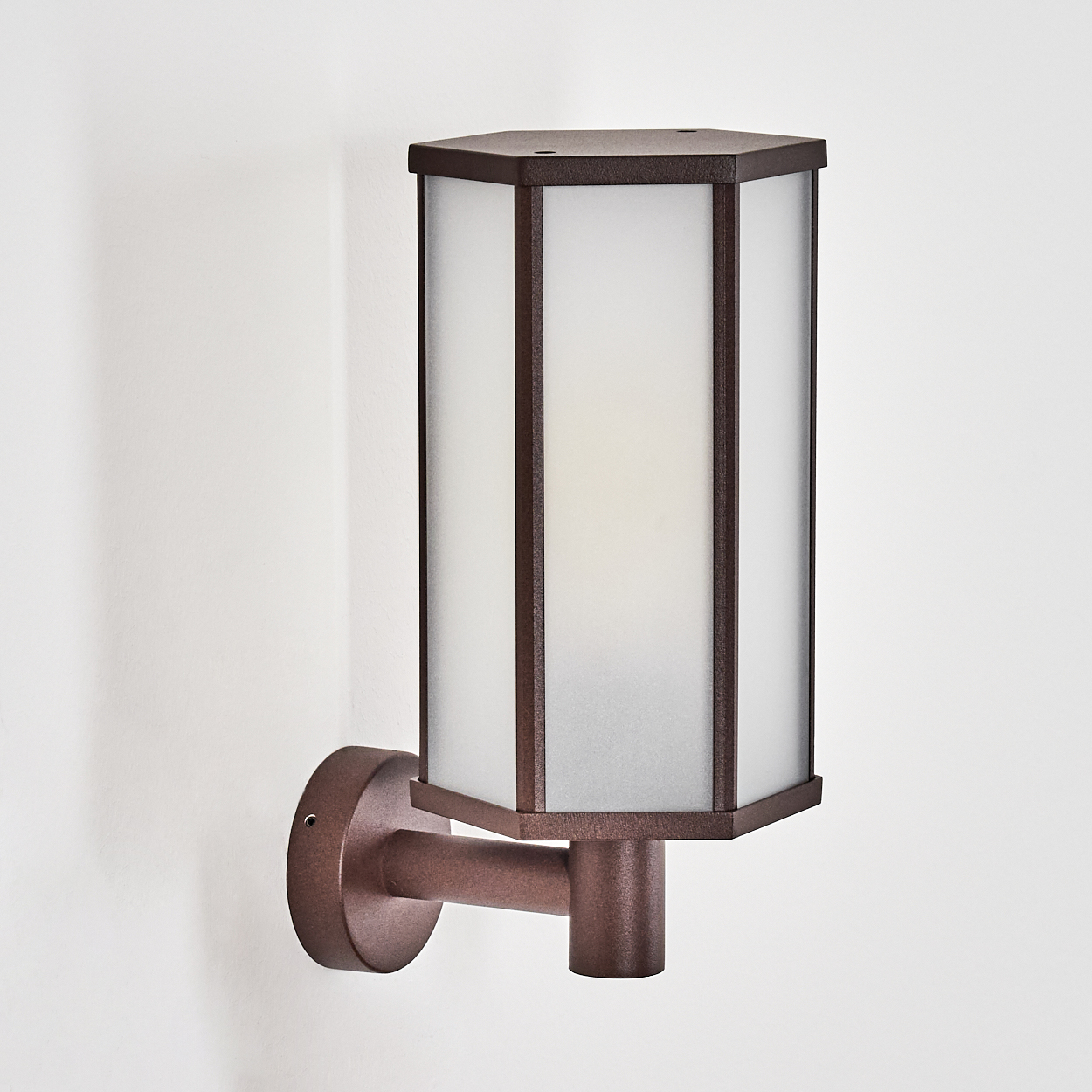 Eine bronzefarbene Wandlampe mit einem Schirm aus Milchglas und sechseckigem Design an einer weißen Wand.