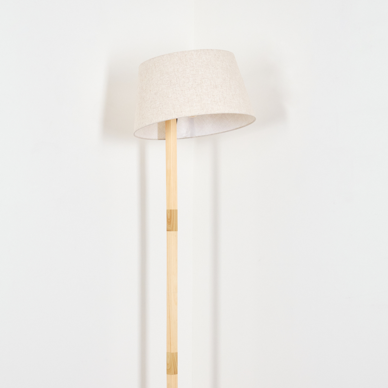 Eine Stehlampe vor einer weißen Wand. Sie hat einen beigefarbenen Lampenschirm und einen hellen Holzständer. Der Hintergrund ist weiß.