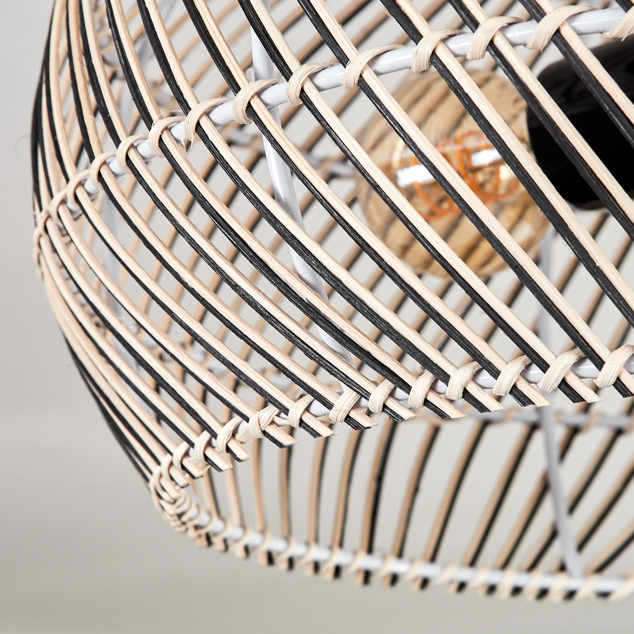 Nahaufnahme eines gewebten Lampenschirms aus Rattan mit schwarzen und naturfarbenen Streifen, weißem Metallrahmen und einer sichtbaren Glühbirne.