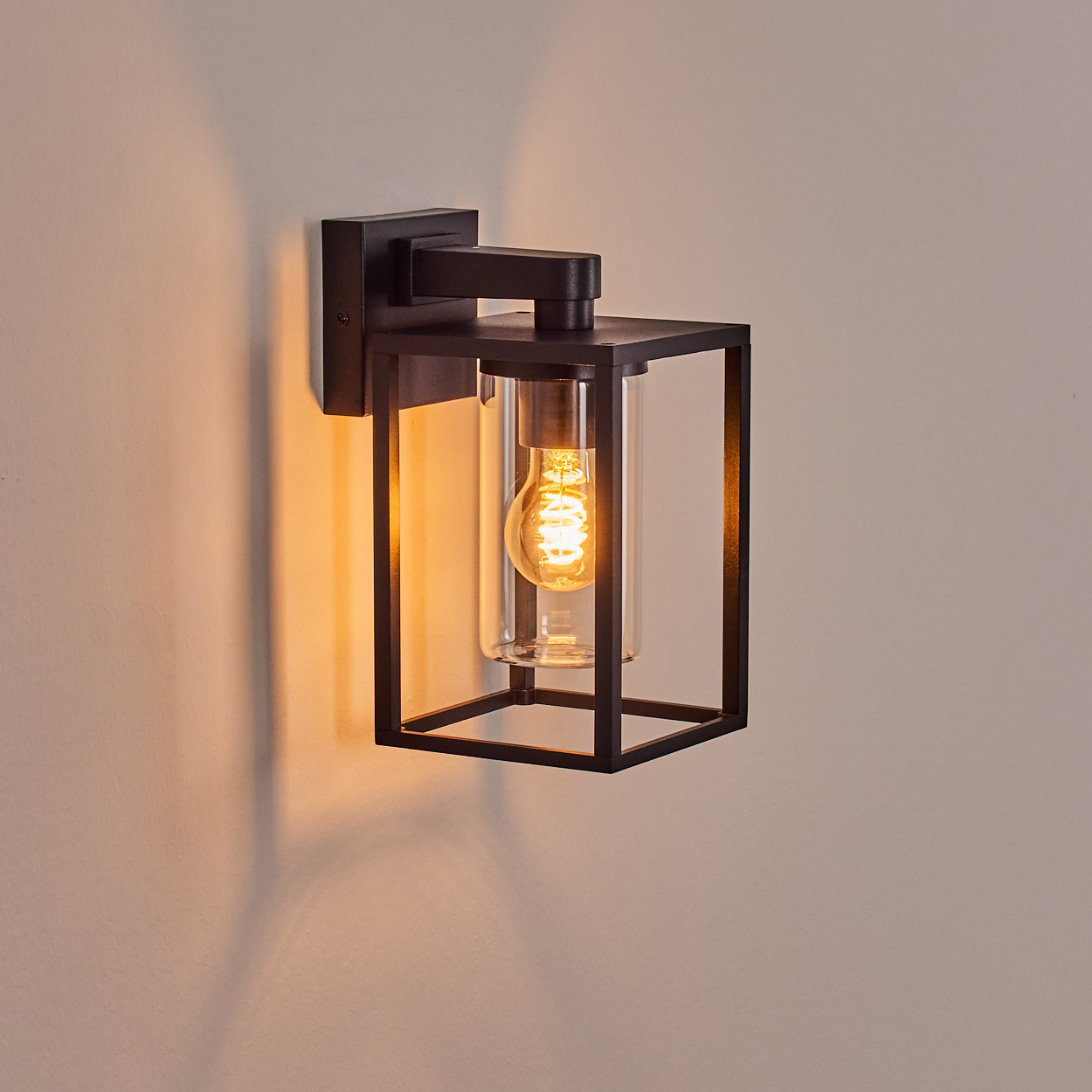 Eine schwarze, rechteckige Außenwandleuchte mit Glasgehäuse, in dem eine leuchtende Edison-Glühbirne untergebracht ist, an einer beigen Wand befestigt.