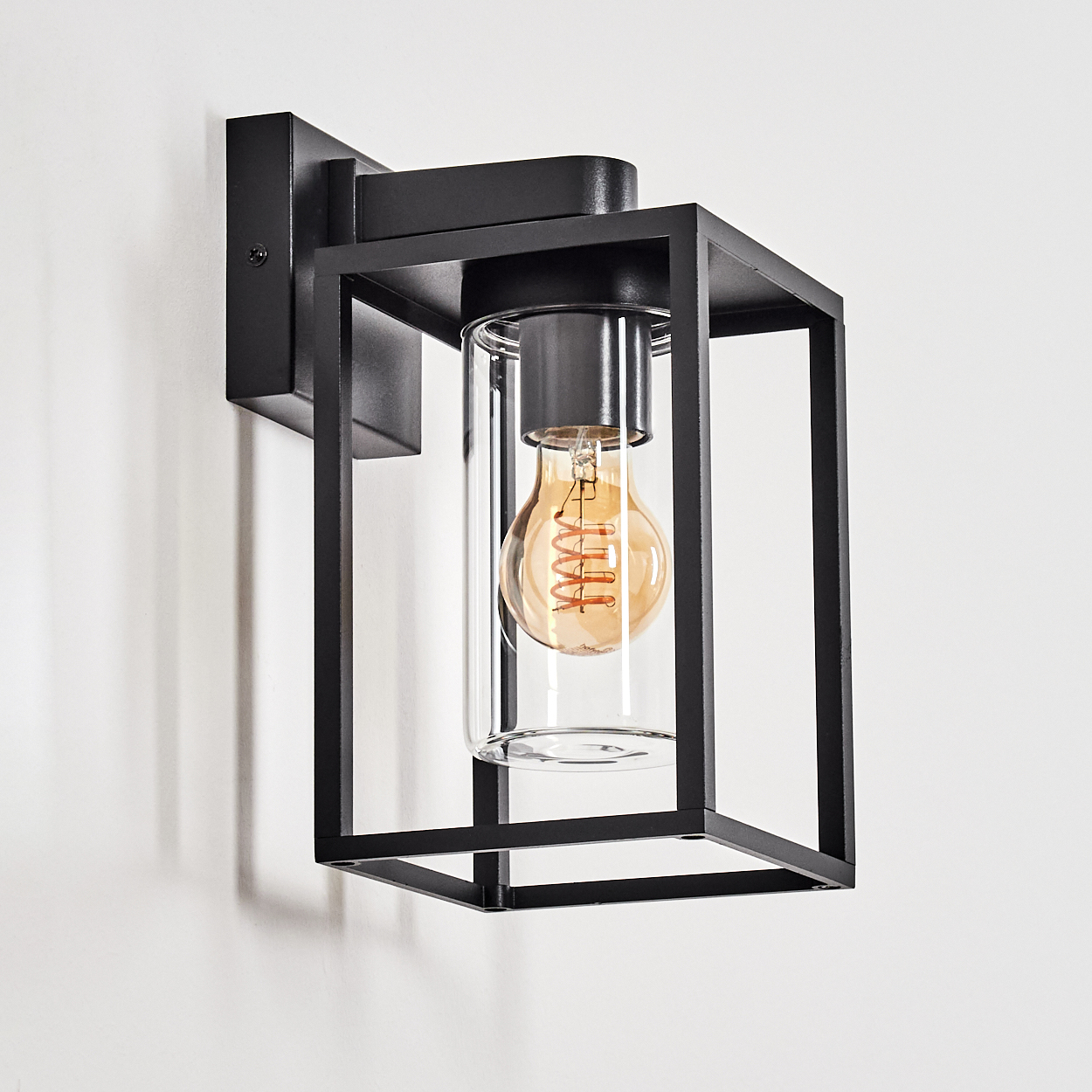 Schwarze Metall-Außenwandleuchte mit Glaskylinder und Glühbirne, montiert an einer weißen Wand. Die Lampe hat ein rechteckiges Design.