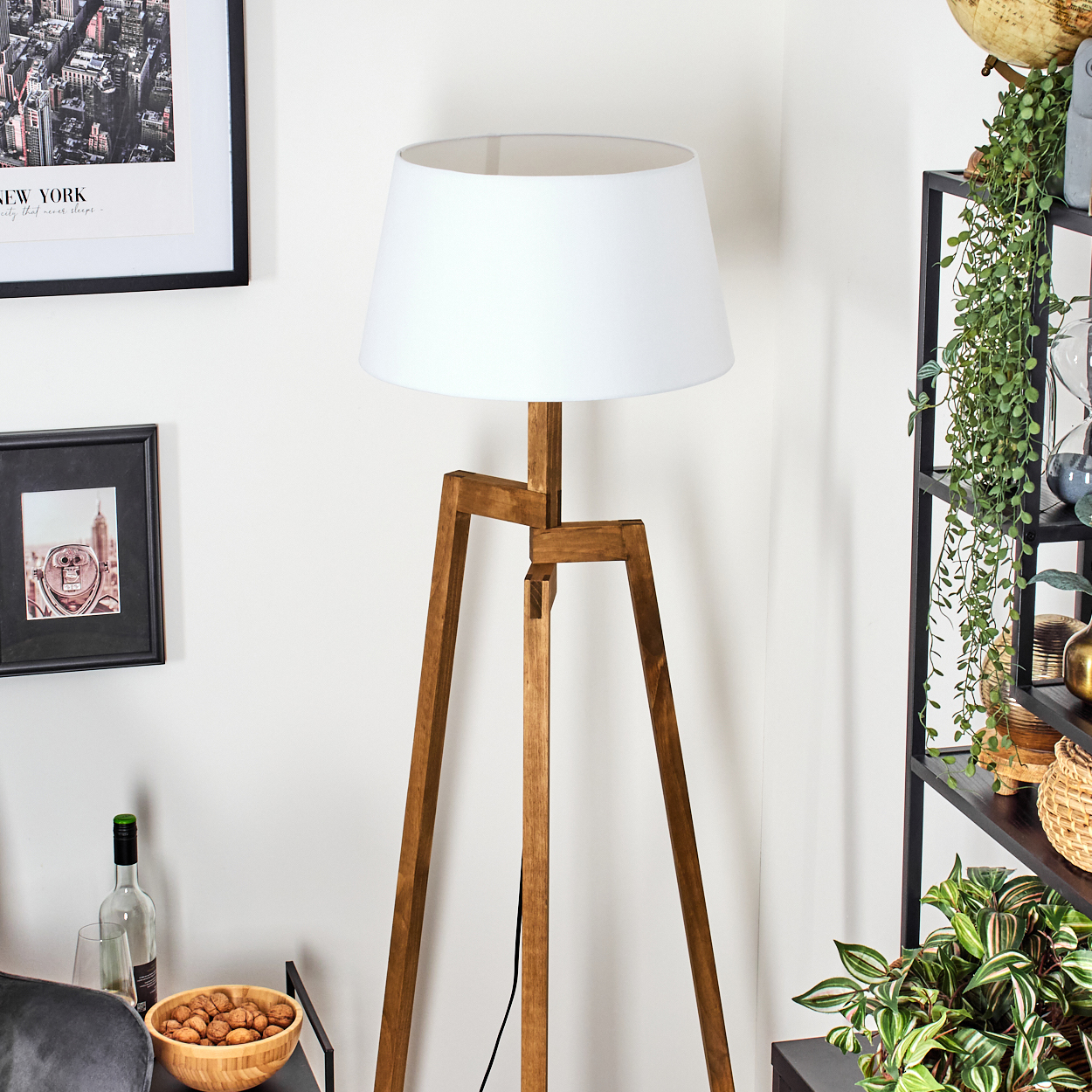 Eine hölzerne Stativ-Stehlampe mit weißem Lampenschirm steht in einem Raum. Im Hintergrund befinden sich Bilder, Pflanzen und Regale.