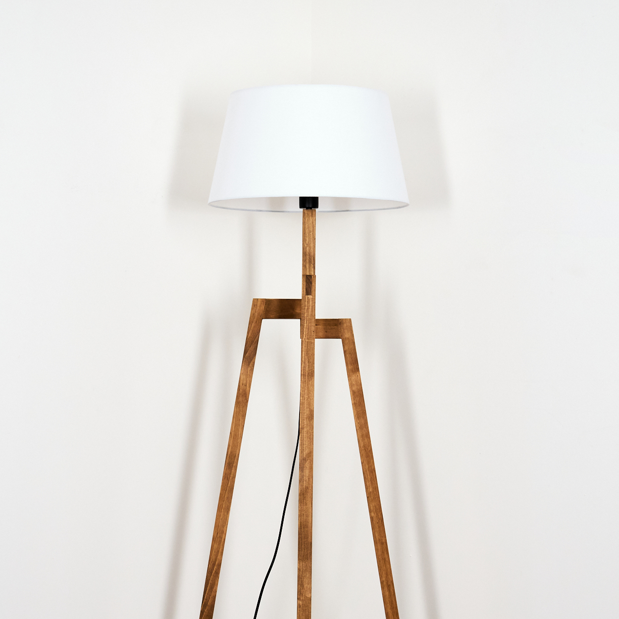 Eine hölzerne Stehlampe mit weißem Lampenschirm steht vor einer weißen Wand. Ein schwarzes Kabel hängt herunter.