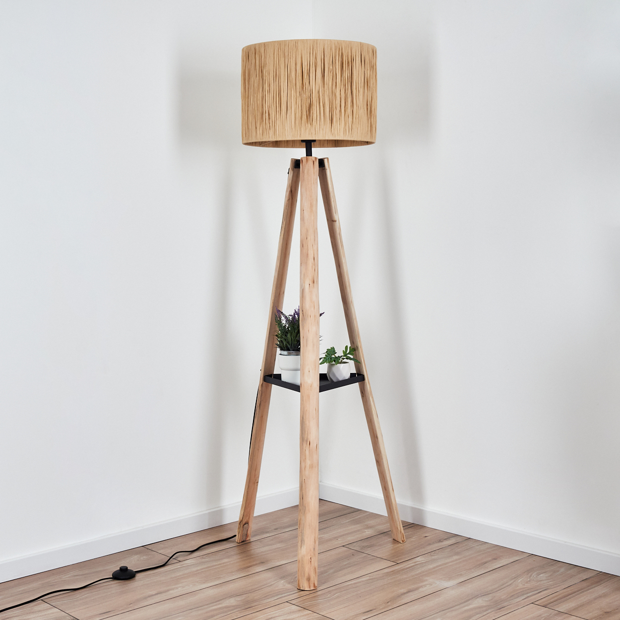 Eine hölzerne Stativ-Stehlampe mit beigem Schirm. Sie hat ein schwarzes Regal mit Topfpflanzen. Der Boden ist aus Holz.