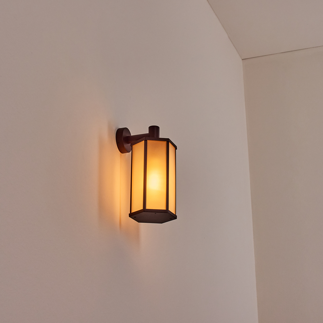 Eine beleuchtete, dunkelbraune Wandleuchte mit rechteckigem, mattiertem Glasschirm ist an einer beigen Wand montiert.