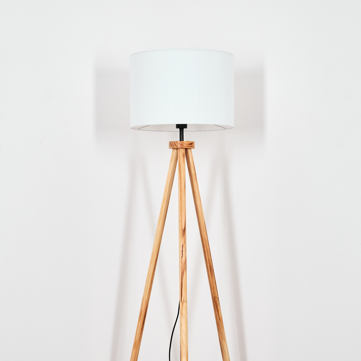 Eine hölzerne Stehlampe mit weißem Lampenschirm steht vor einer weißen Wand. Ein schwarzes Kabel ist sichtbar.