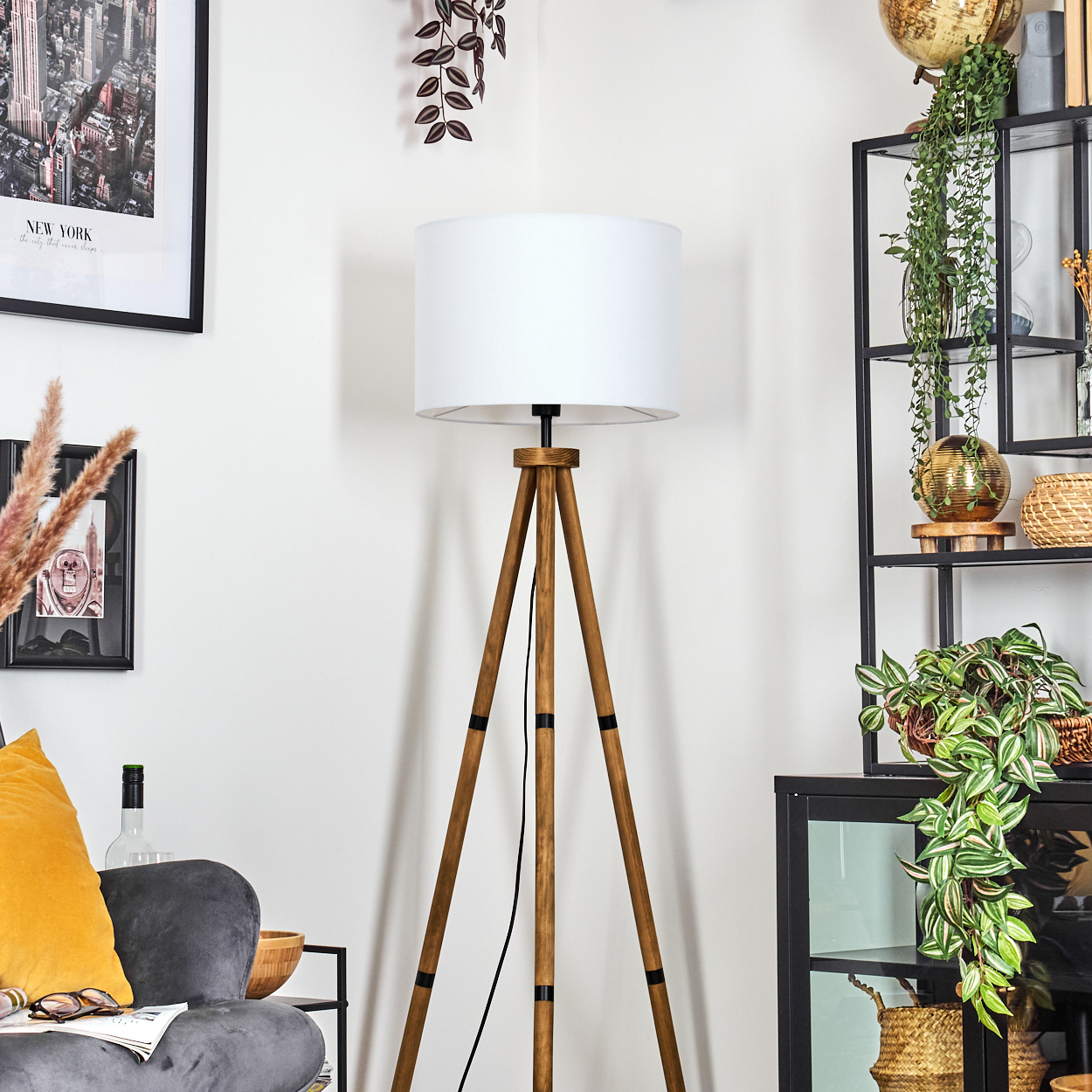 Eine Holz-Stativlampe mit weißem Schirm steht in einer Ecke. Ein grauer Sessel mit gelbem Kissen steht daneben. Regale mit Pflanzen.