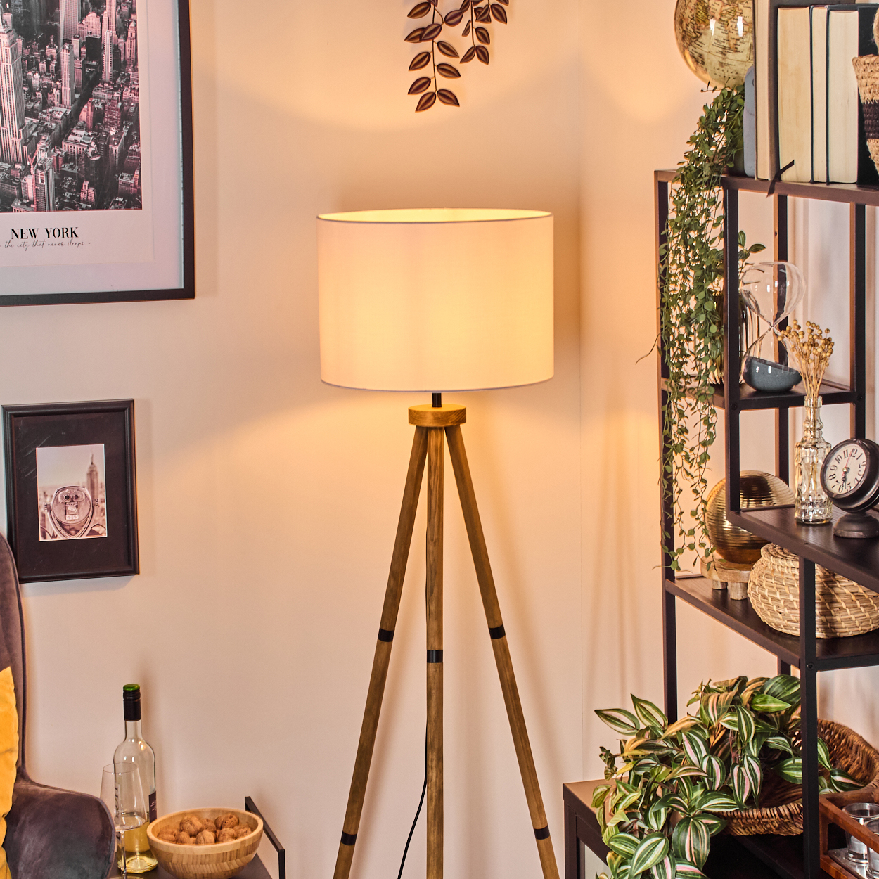 Eine Holz-Stativlampe mit weißem Lampenschirm steht in einem Raum. Regale sind mit Büchern, Pflanzen und Dekorationsartikeln gefüllt.