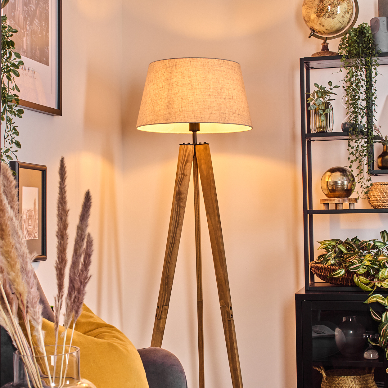Eine Holz-Dreibein-Lampe mit beigem Schirm steht in einem Raum. Regale mit Pflanzen und Dekoration befinden sich rechts. Links ein Sofa und Kunst.