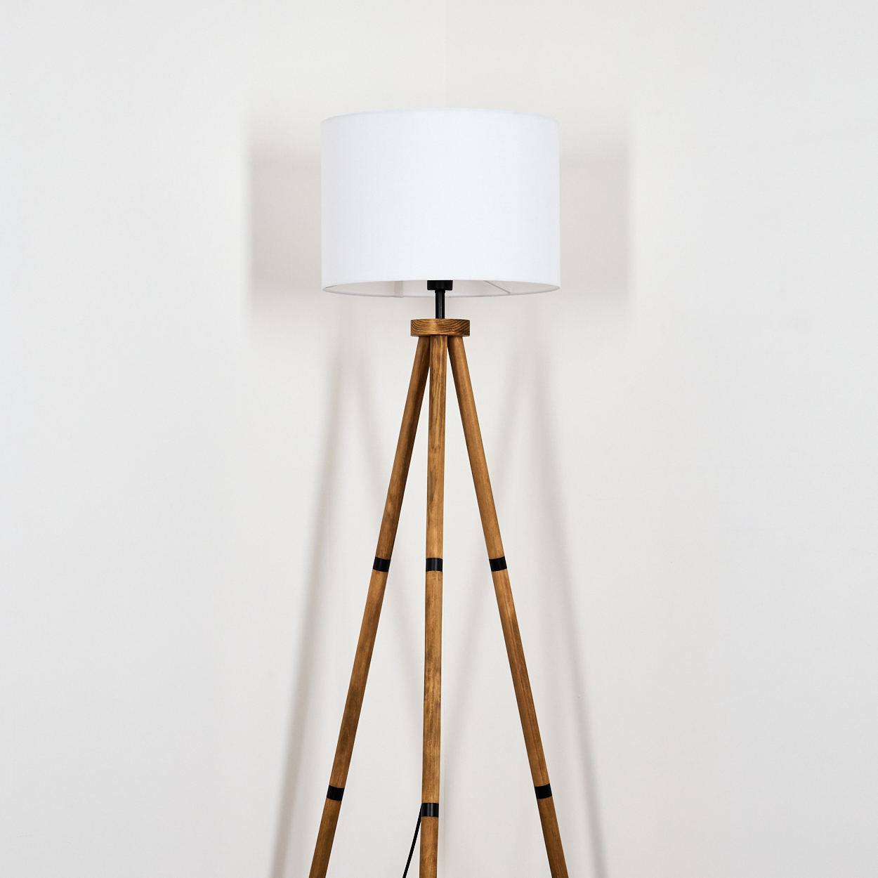 Eine Stehlampe mit Holzstativ und weißem Lampenschirm steht vor einer weißen Wand. Schwarze Akzente an den Beinen und ein schwarzes Kabel sind sichtbar.