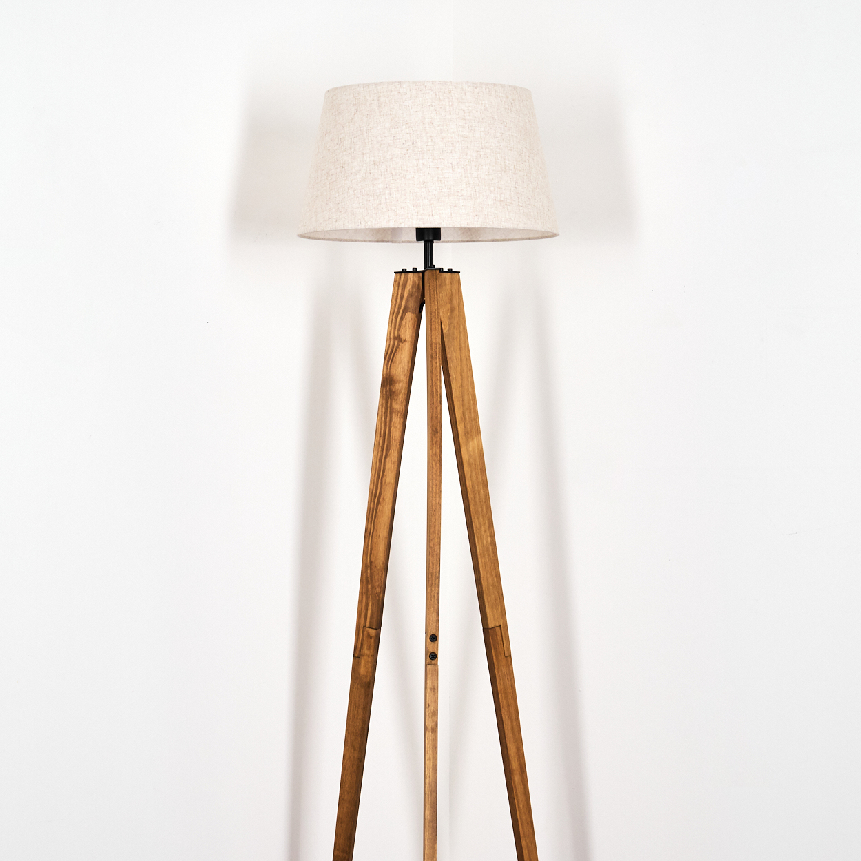 Eine hölzerne Stativ-Stehlampe mit beigem Lampenschirm steht vor einer weißen Wand. Die Holzbeine sind braun.